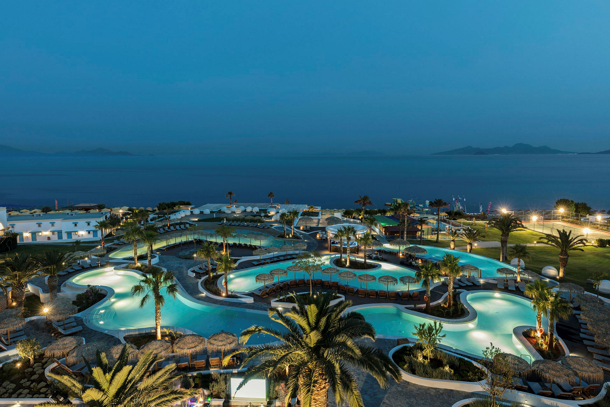 Utsikt över ett upplyst hotellområde med pooler och palmer under kvällstid, med havet i bakgrunden. (AI-genererad text)