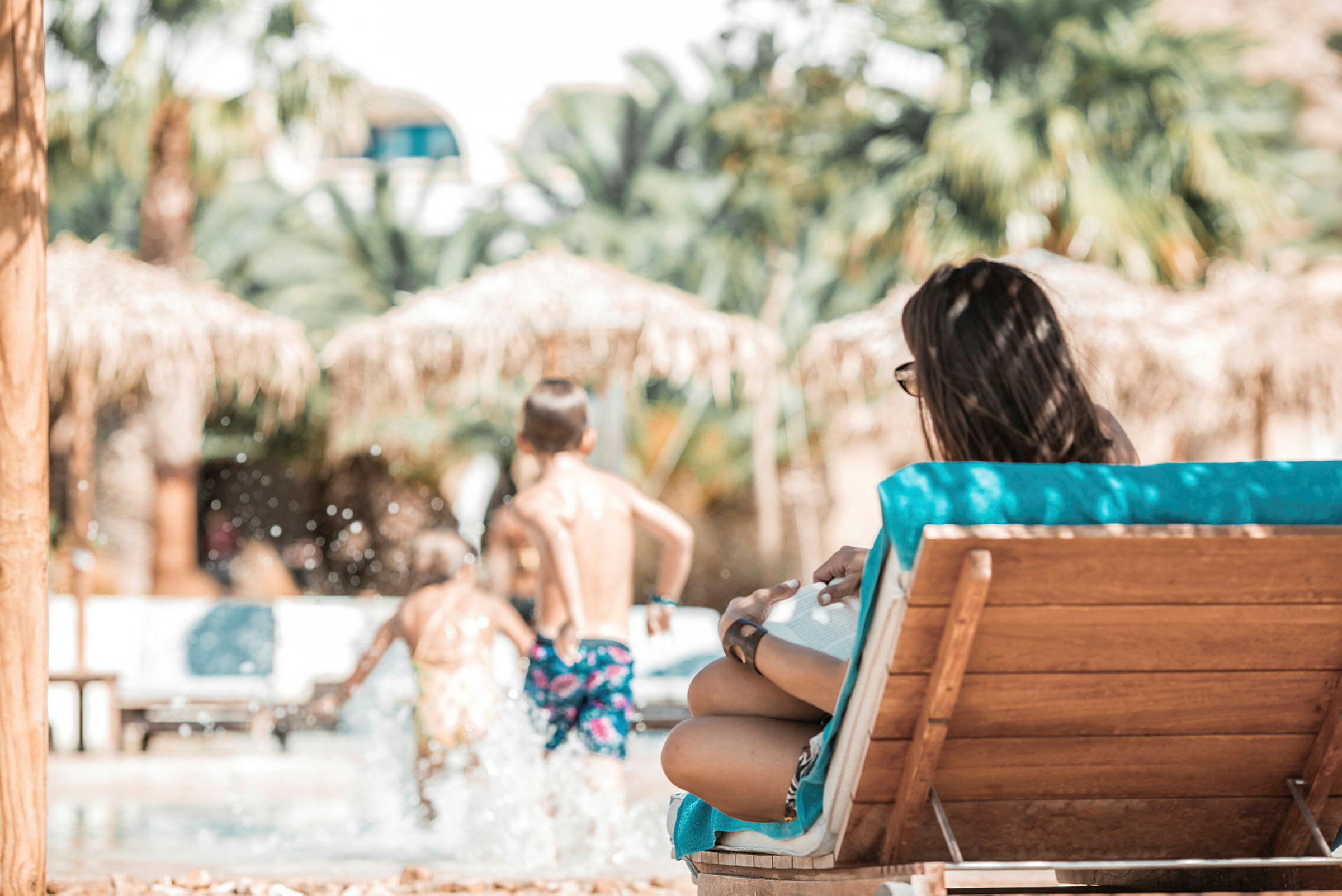 En person sitter på en solstol och läser nära en pool där två barn leker. (AI-genererad text)