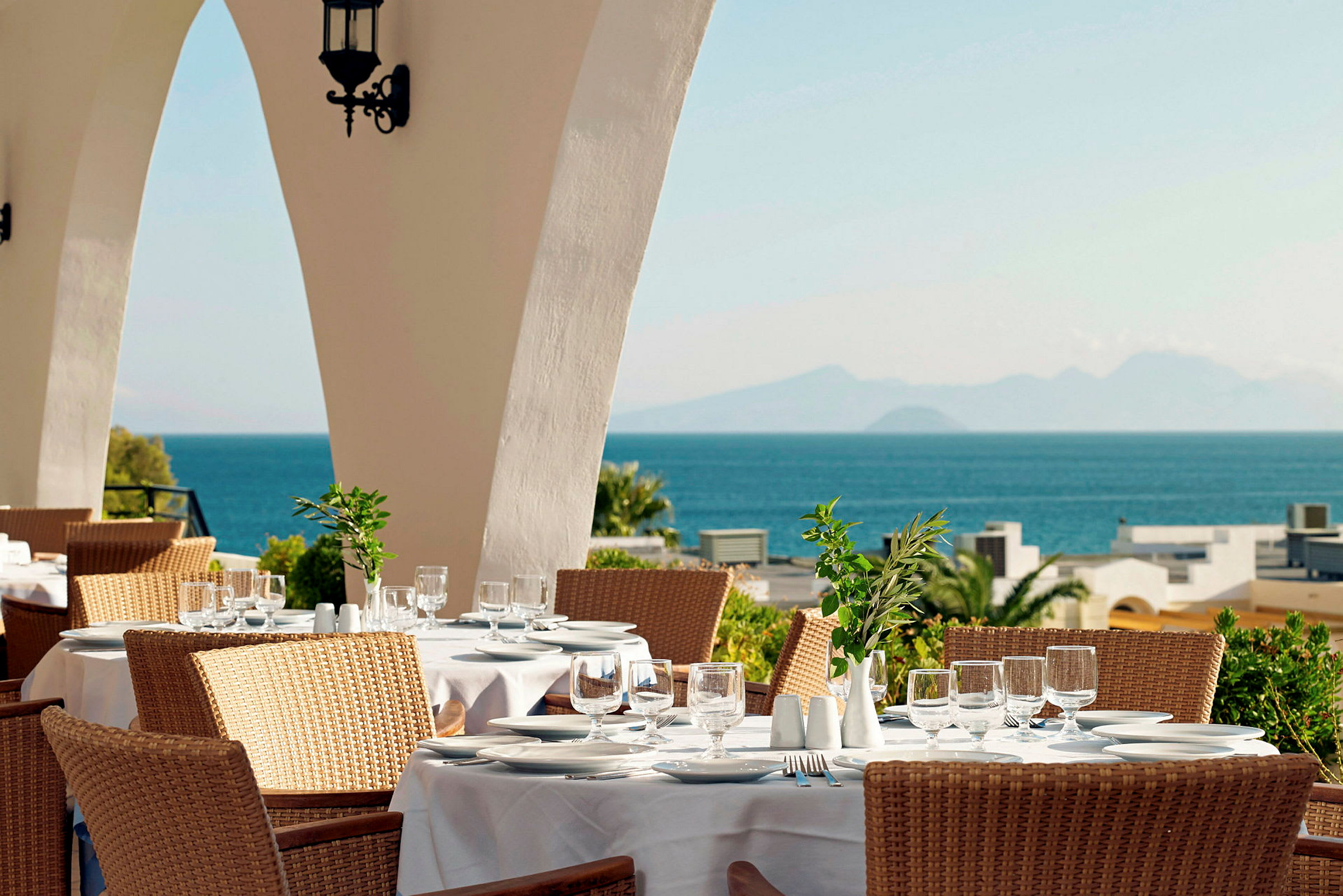 En restaurang med bord dukade med vita dukar, en solig utsikt över havet och berg i bakgrunden. (AI-genererad text)