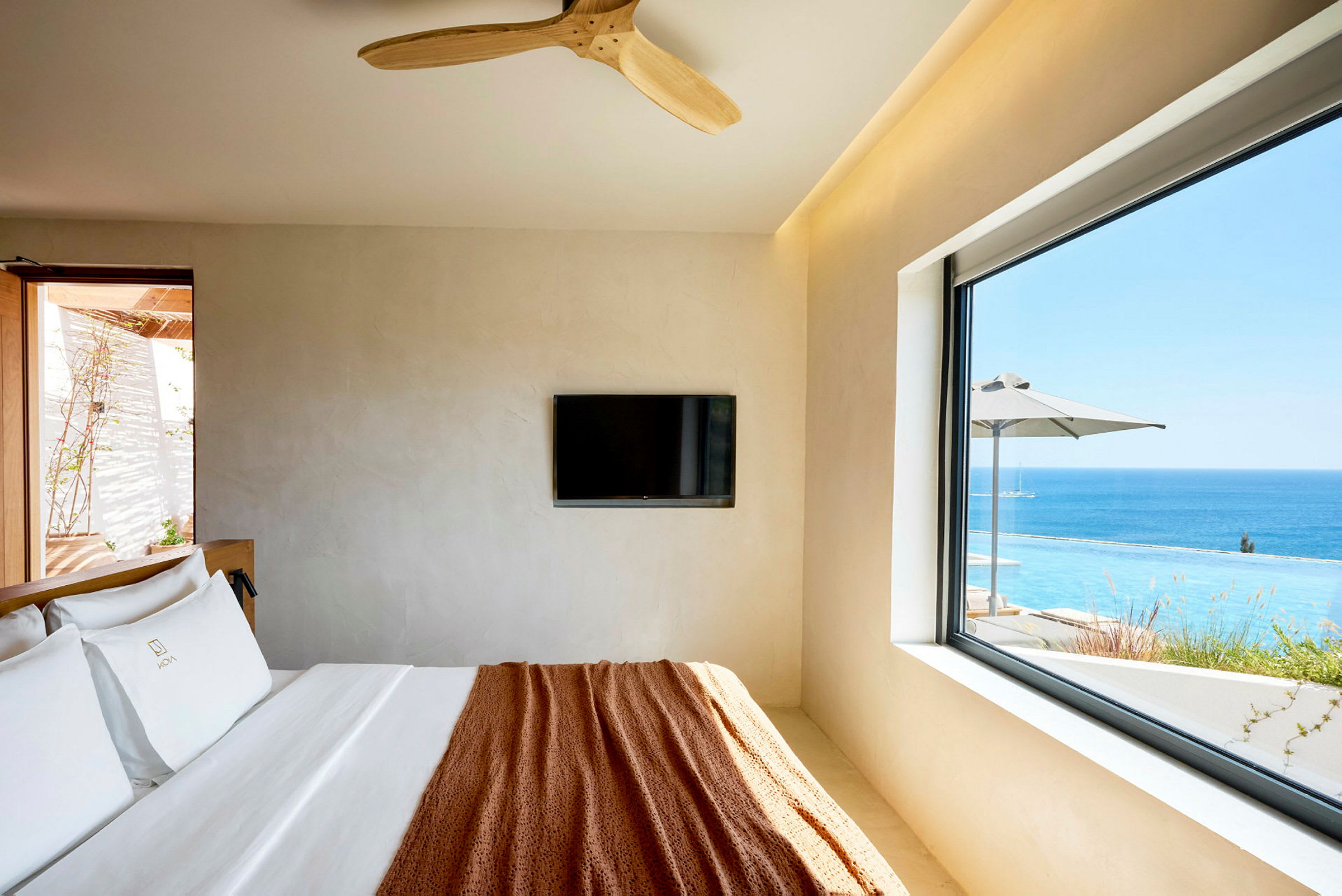 Ett hotellrum med en säng, väggmonterad TV och fönster med utsikt över en pool och havet. (AI-genererad text)