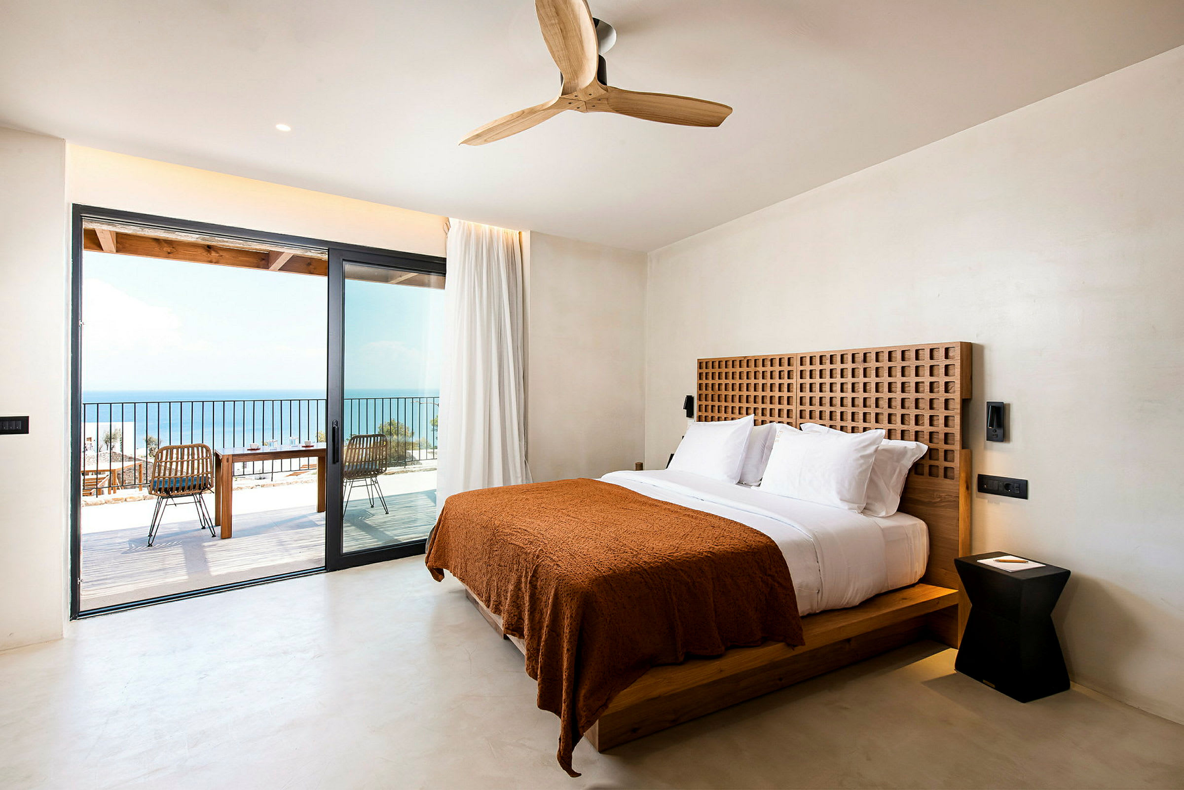 Ett modernt hotellrum med en dubbelsäng med vita sängkläder och en brun filt. Rummet har en stor glasdörr som leder till en balkong med utsikt över havet. (AI-genererad text)