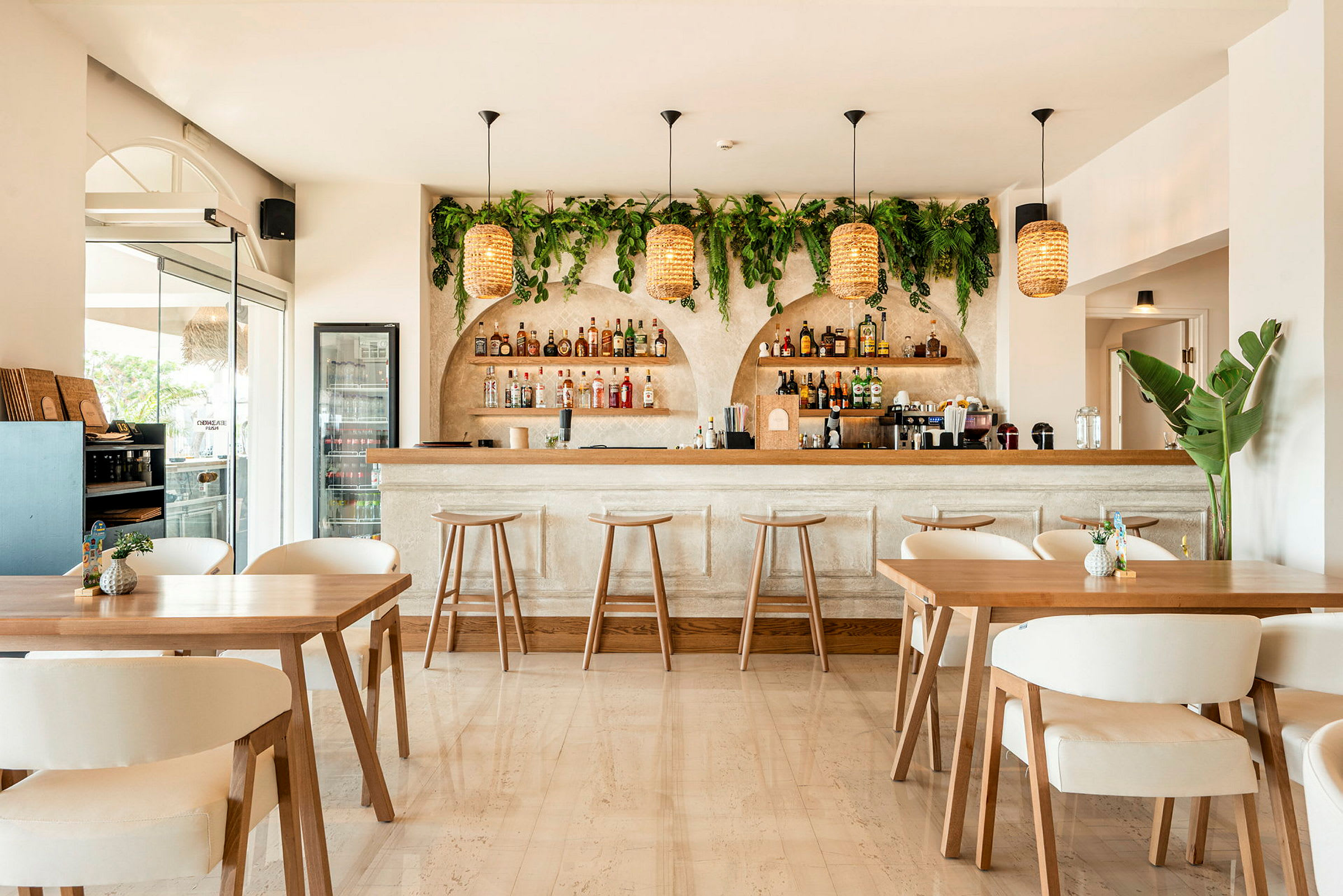 Bild på en stilren bar med ljus inredning. Baren har omslutande grön växtdekoration samt hängande flätade lampor. (AI-genererad text)