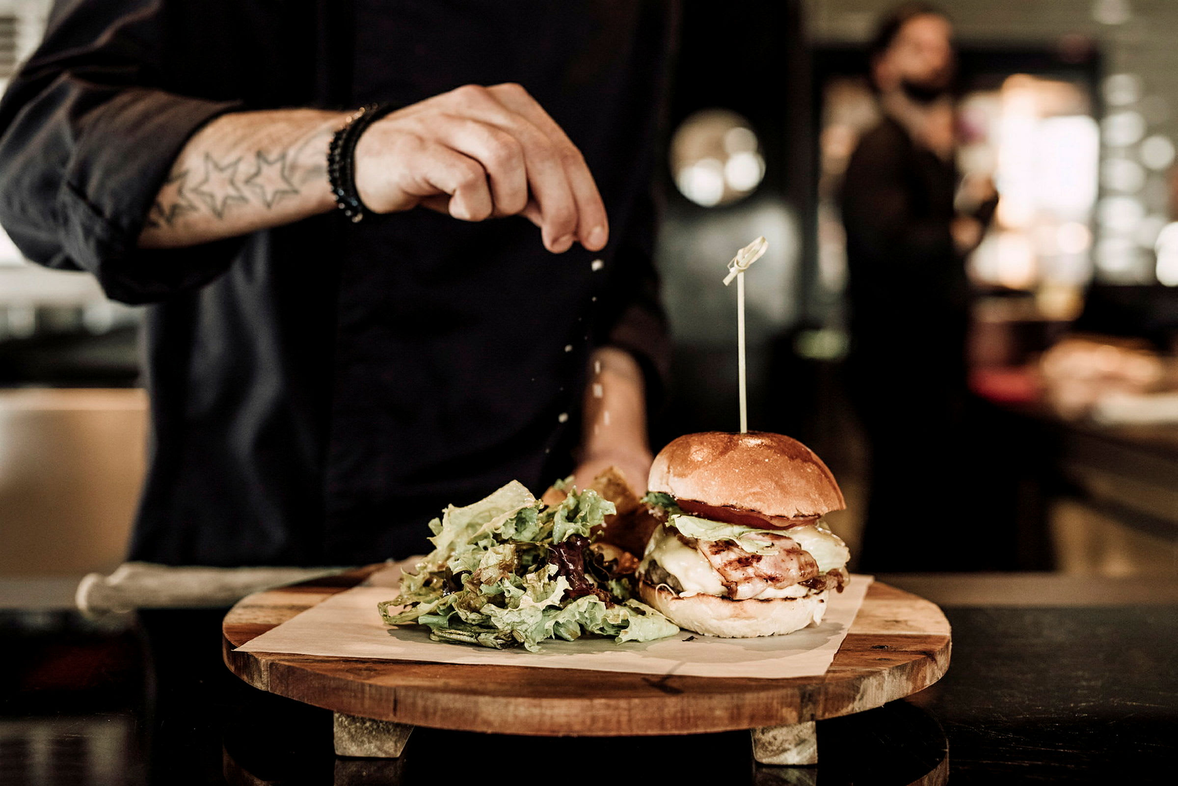 En person kryddar en hamburgare som är placerad på en träplatta tillsammans med en portion sallad. (AI-genererad text)
