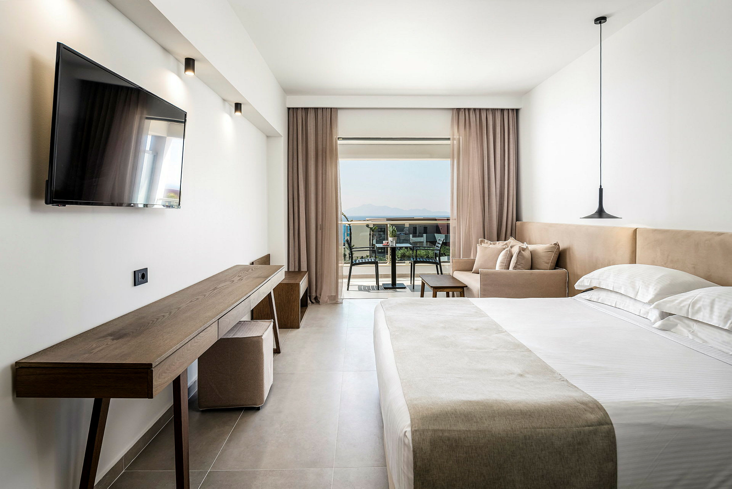 Interiör av ett hotellrum med en dubbelsäng, soffa och skrivbord. Bakom glasdörrar syns en möblerad balkong med utsikt över omgivningen. (AI-genererad text)