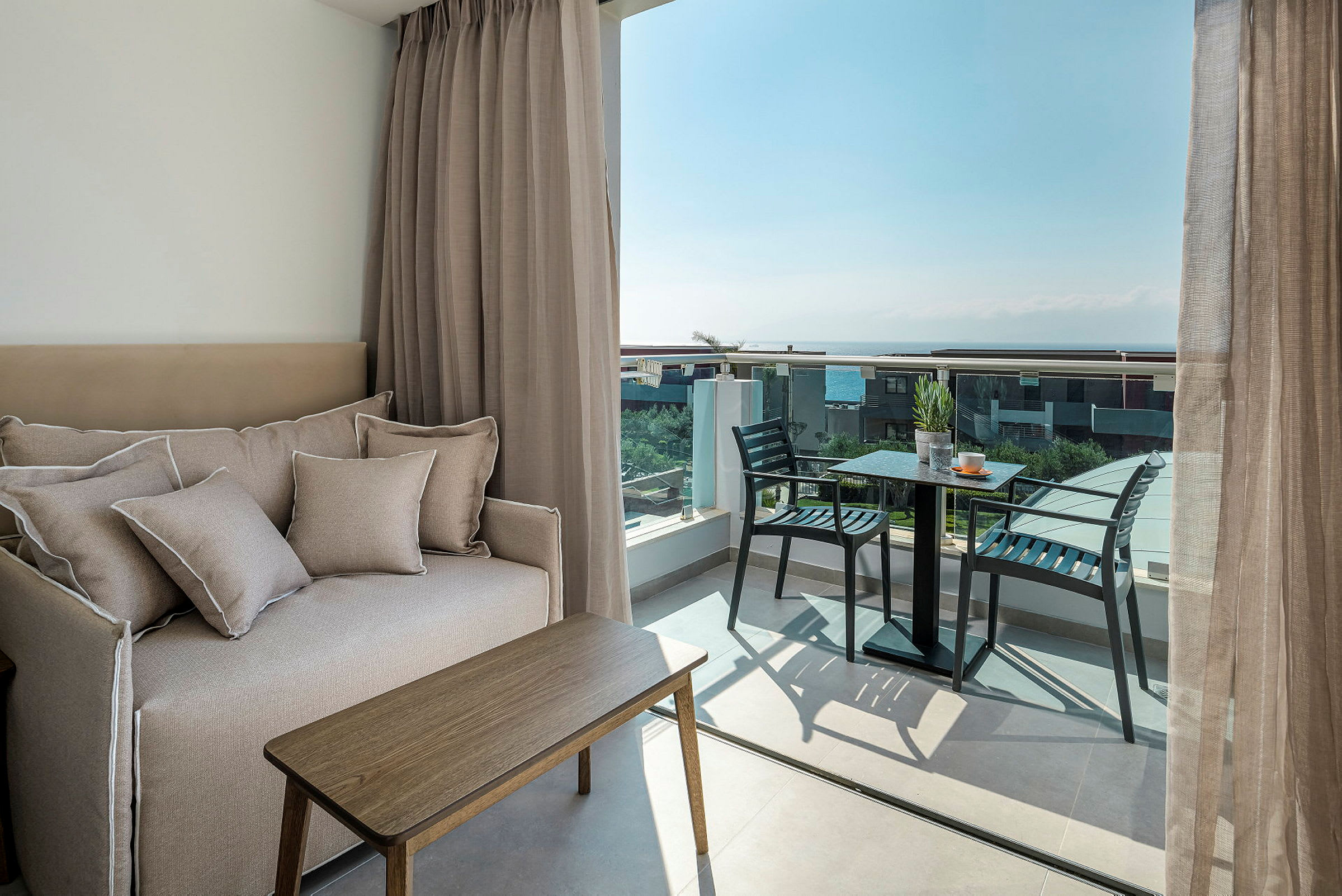 Bild av ett hotellrum med en soffa, ett litet bord och en balkong med stolar och bord samt utsikt över havet. (AI-genererad text)