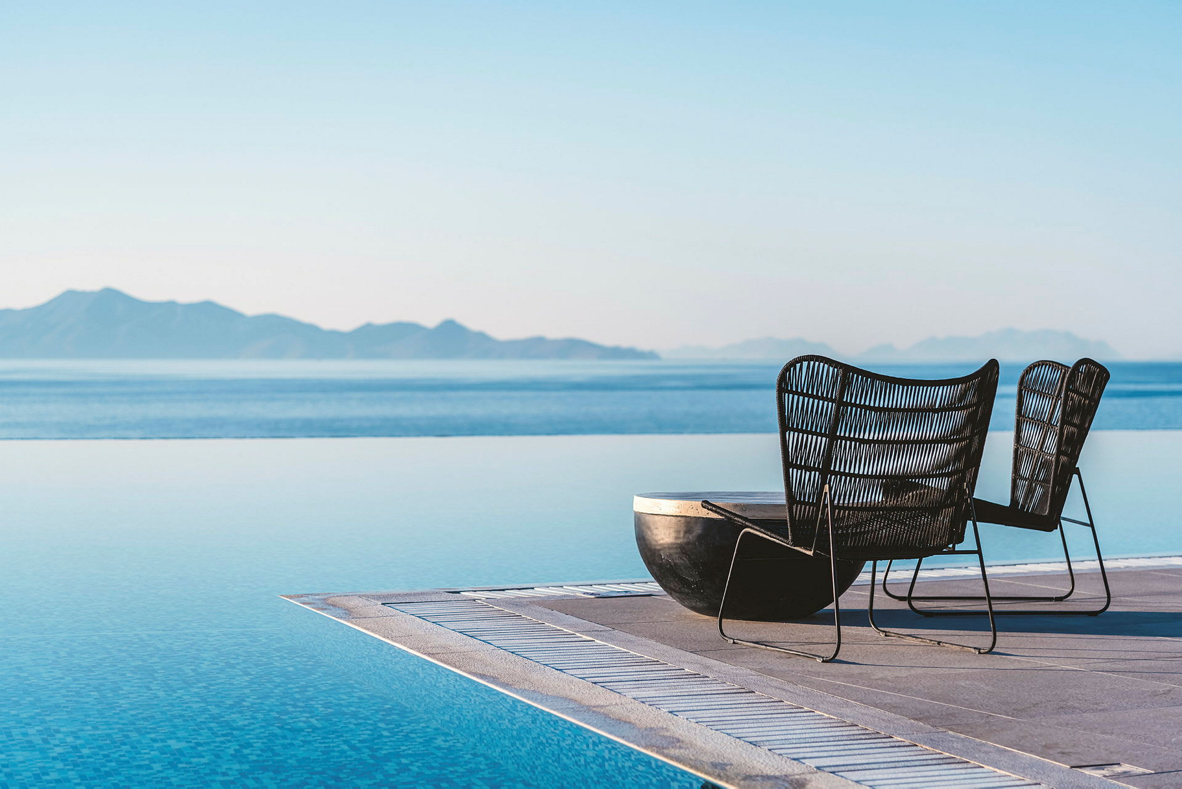 Två stolar vid kanten av en infinitypool med hav och berg i bakgrunden. (AI-genererad text)