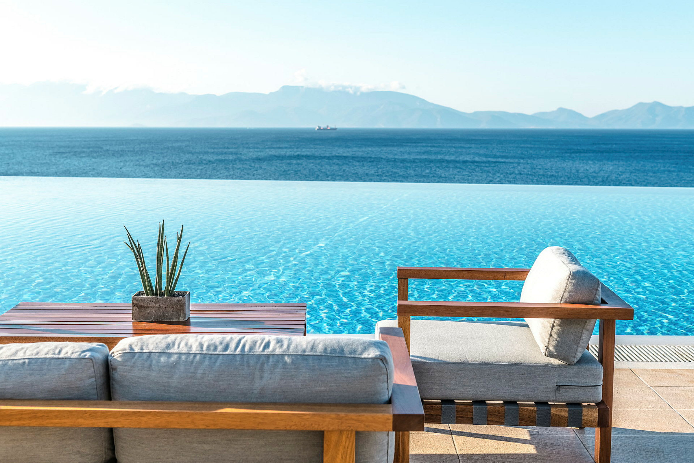 En infinitypool med utsikt över en blå hav och avlägsna berg, med trämöbler och en liten växt på ett bord i förgrunden. (AI-genererad text)
