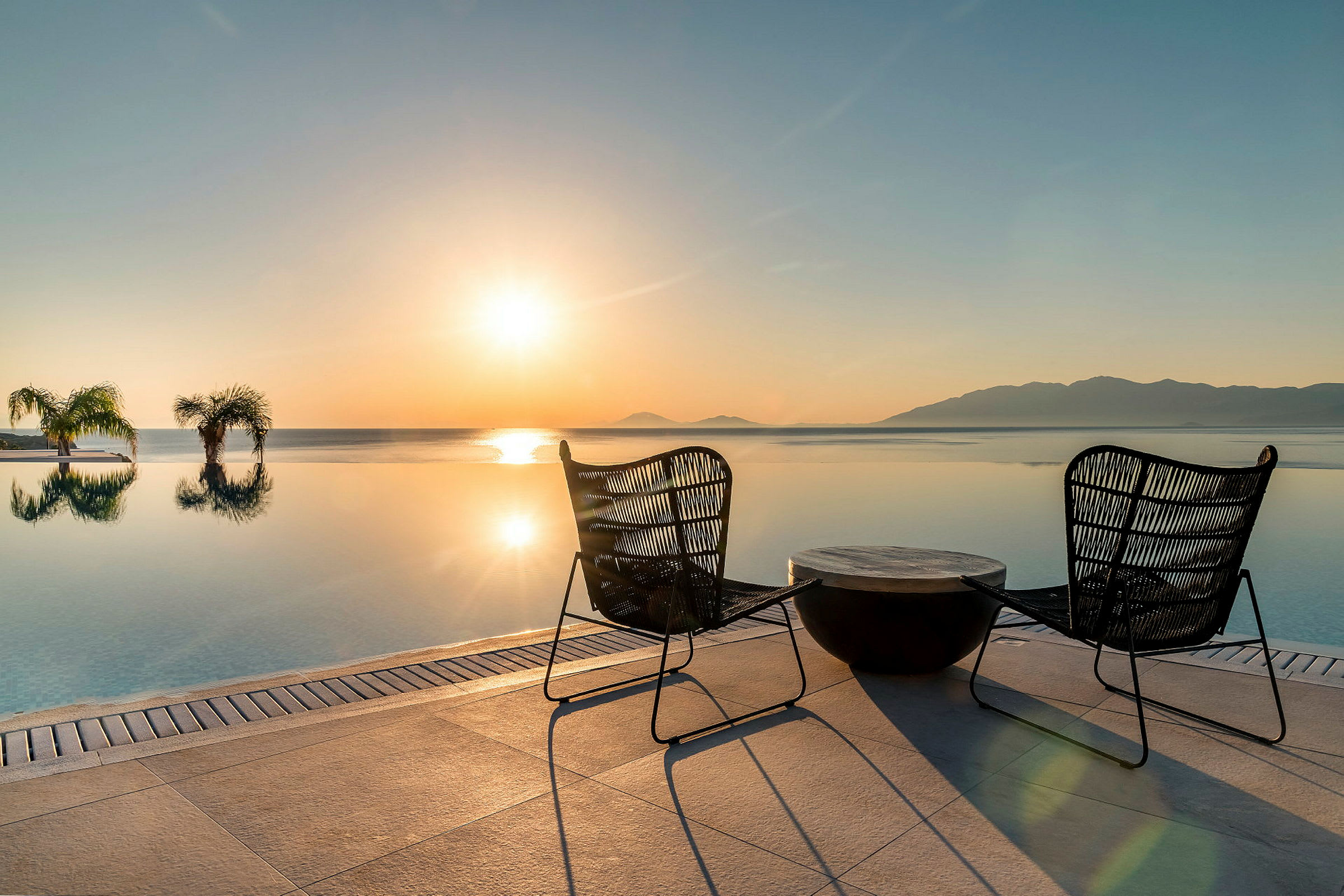 Två stolar och ett bord vid en infinitypool med utsikt över havet i solnedgång. (AI-genererad text)