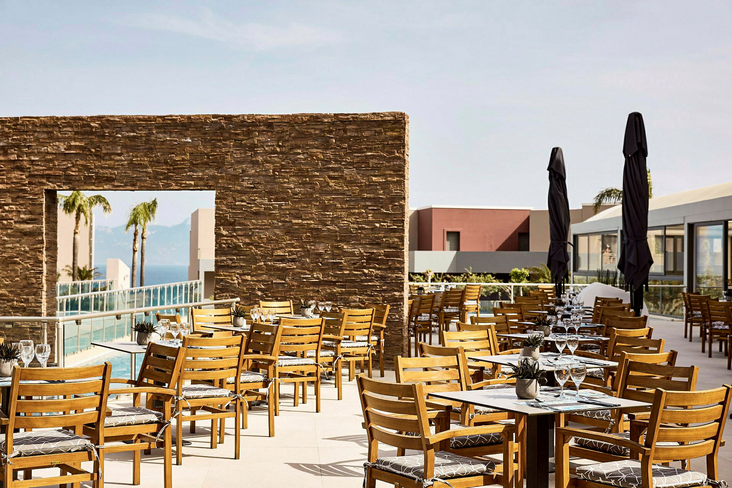 En utomhus restaurang med trästolar och bord, omgiven av en modern byggnad och en pool. (AI-genererad text)