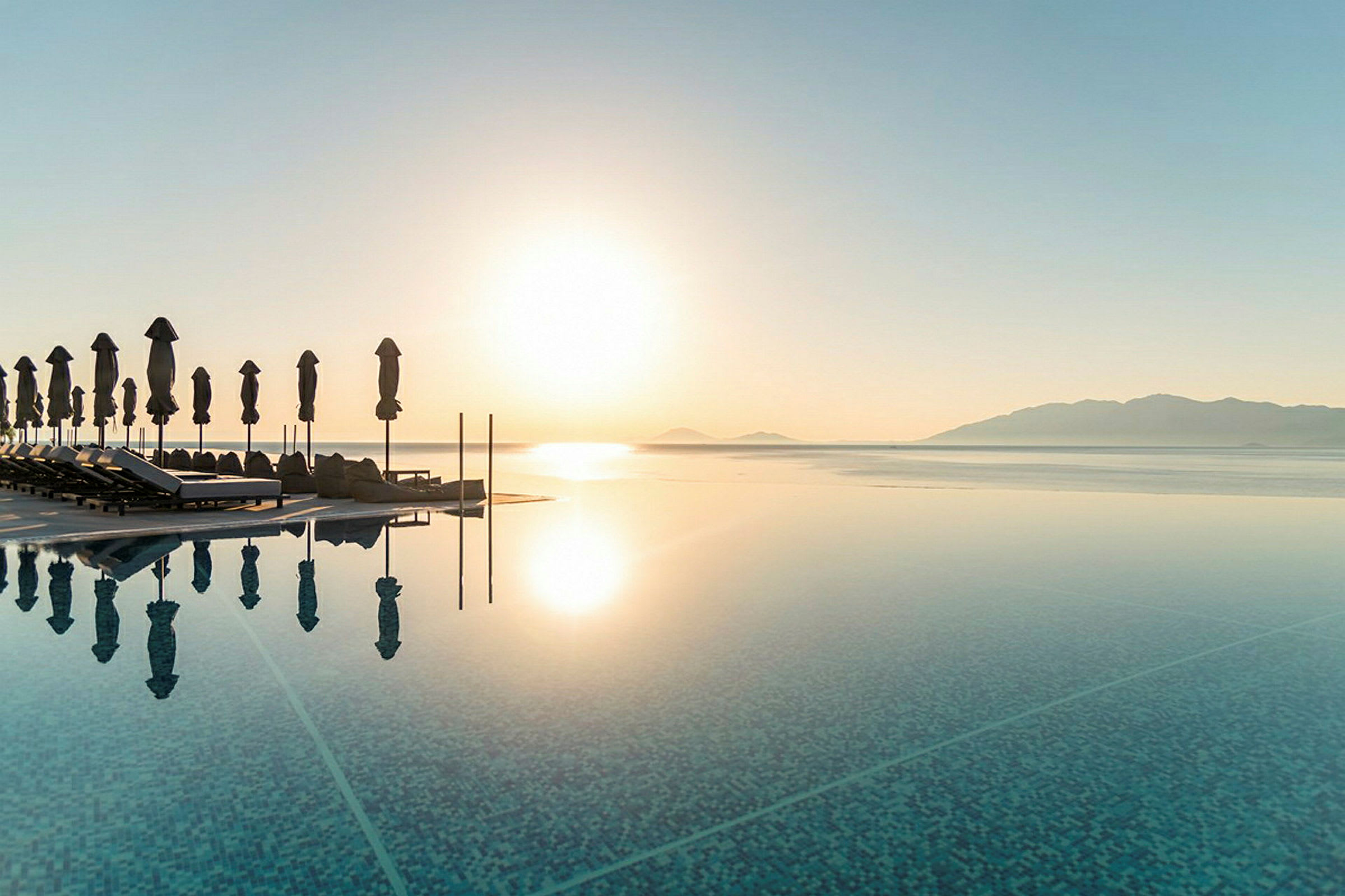 Strand med solstolar och parasoller vid en infinitypool med utsikt över havet och berg i solnedgången. (AI-genererad text)
