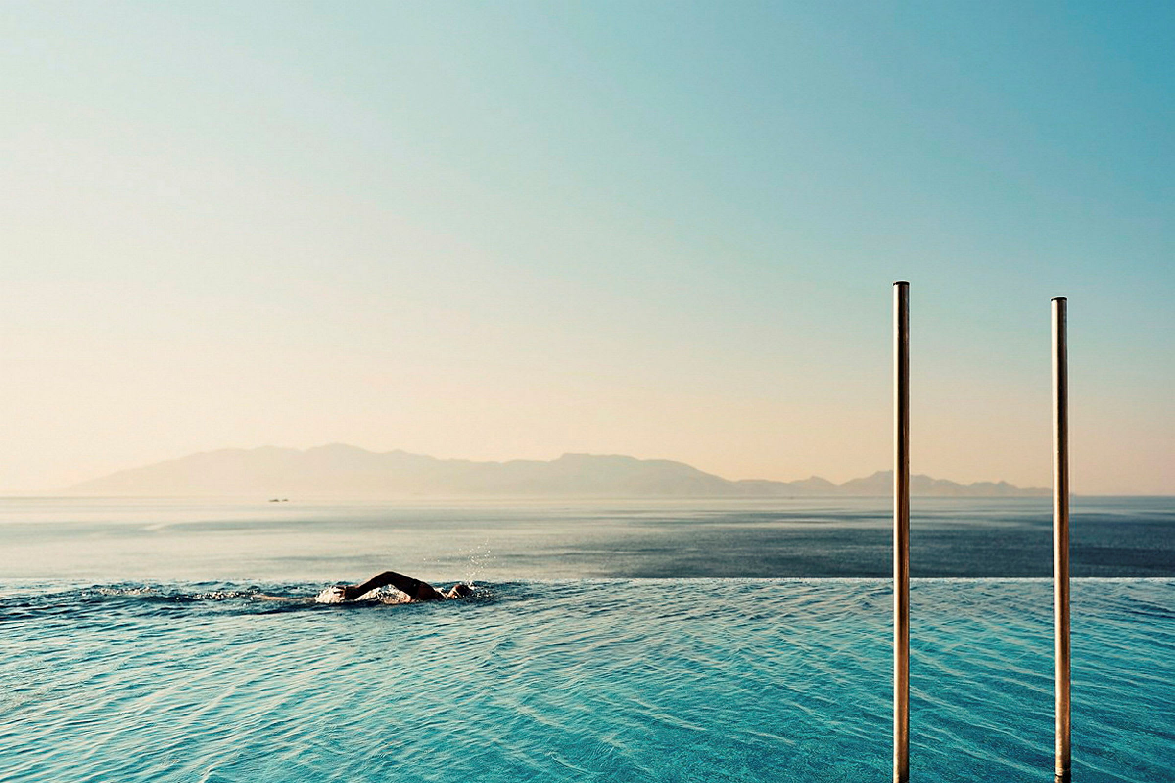 En person simmar i en infinitypool med havet och berg i bakgrunden. (AI-genererad text)