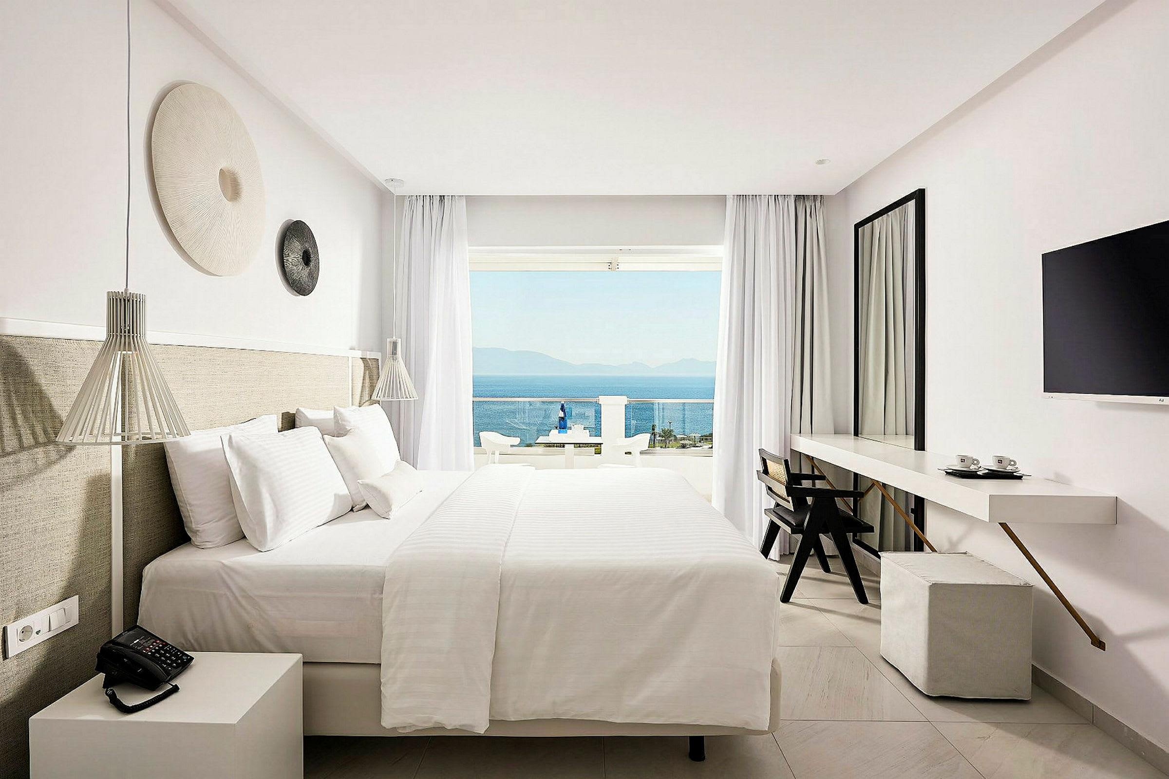 Ett hotellrum med en stor säng, skrivbord, tv och balkong med utsikt över havet. (AI-genererad text)