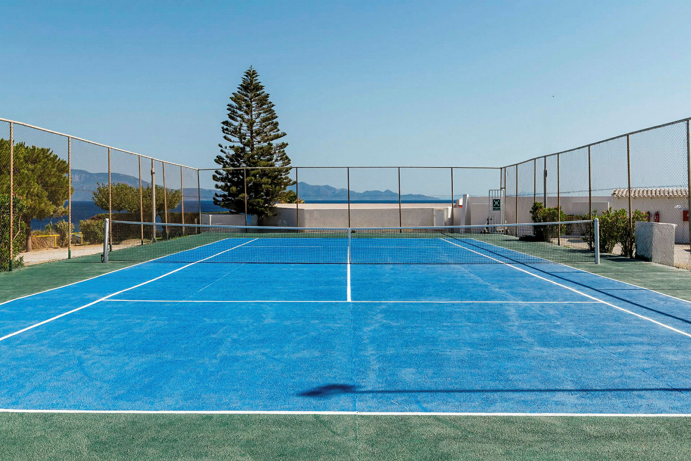 En blå tennisbana vid kusten omgiven av stängsel, med havet och berg i bakgrunden. (AI-genererad text)
