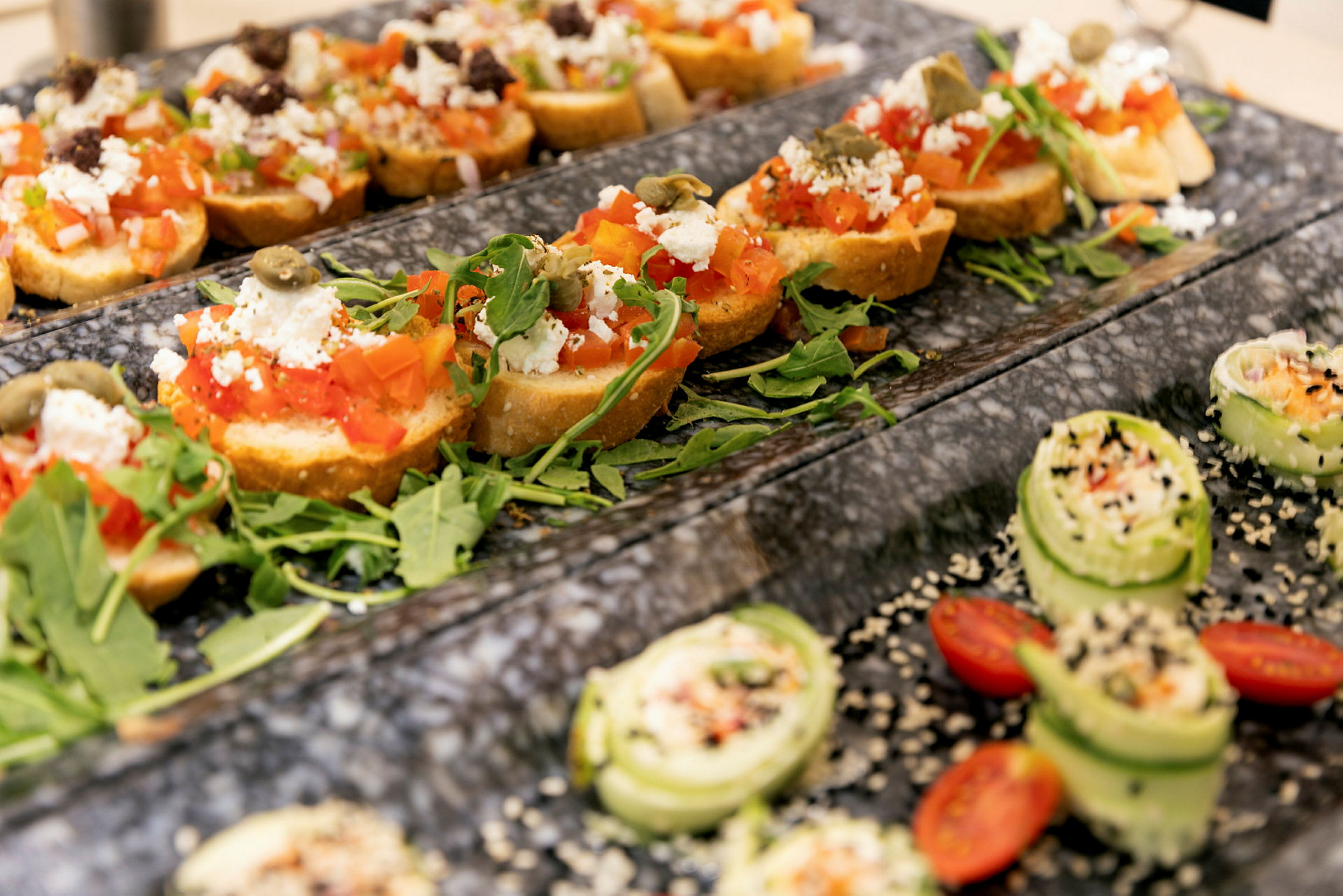 Uppläggning av bruschetta med tomat, fetaost och ruccola samt små gurksnittar med sesamfrön och körsbärstomater. (AI-genererad text)