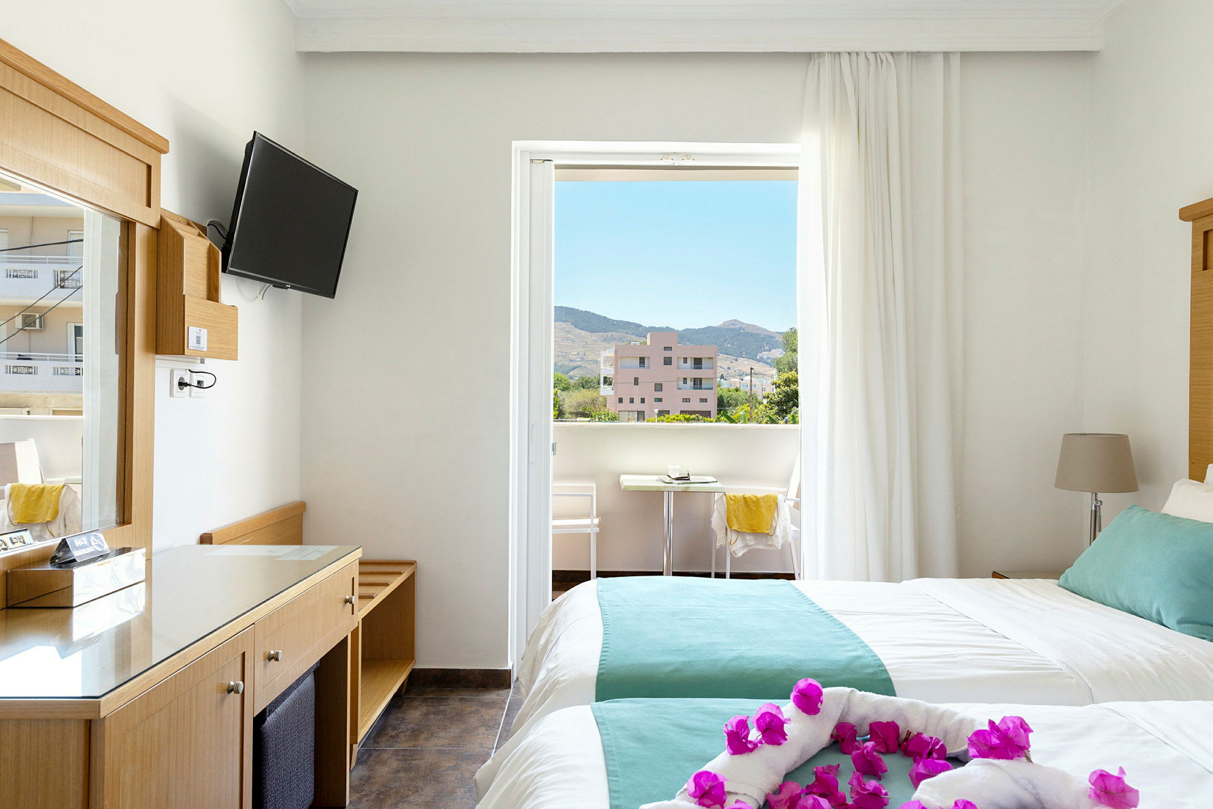 Hotellrum med två bäddar och balkong. Utsikt över omgivande byggnader och berg. (AI-genererad text)