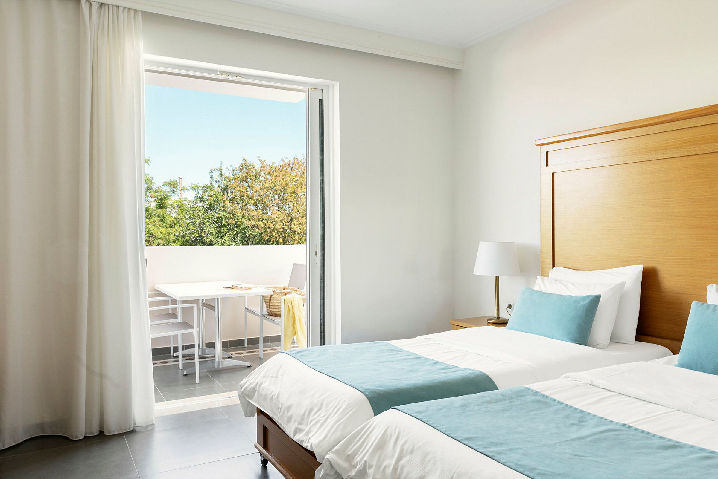 Hotellrum med två enkelsängar, blå sängkläder och en direkt utgång till en balkong med träd som syns i bakgrunden. (AI-genererad text)
