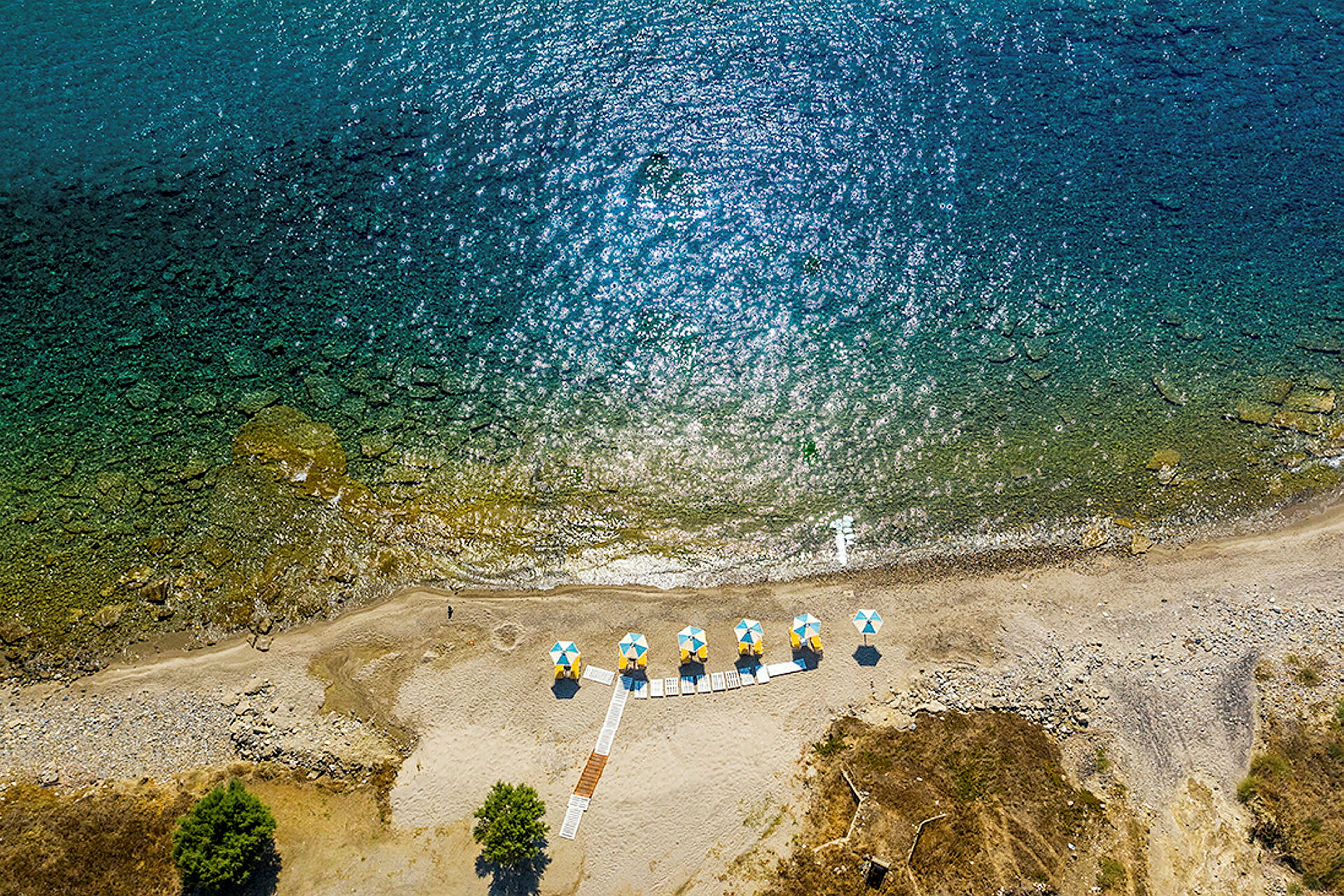 Flygfoto av en strand med parasoller i gula och blå färger samt gångvägar. (AI-genererad text)
