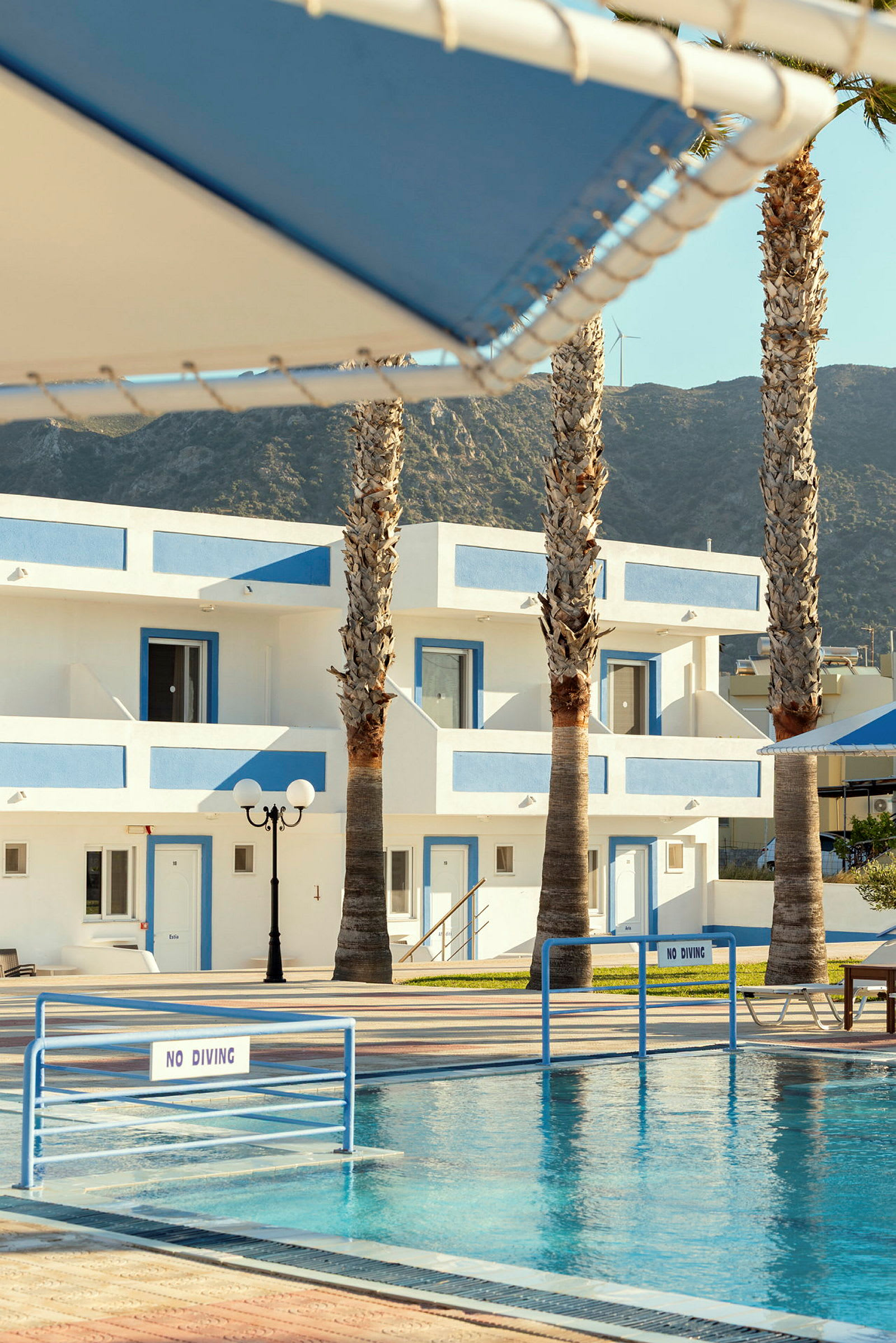 Hotellområde med vit byggnad och blå detaljer, pool och palmer. (AI-genererad text)