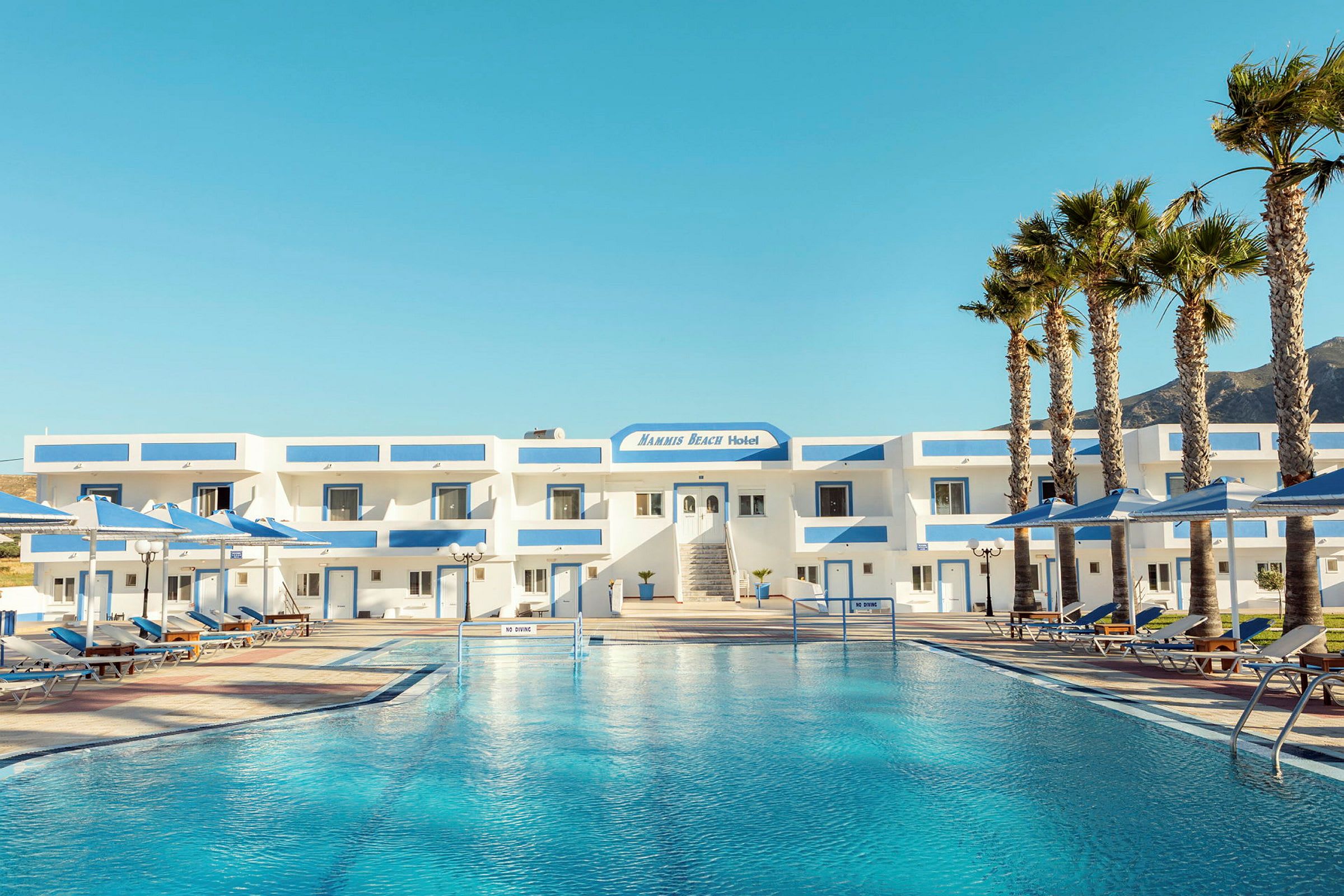 Hotellbyggnad med en pool framför, omgiven av solstolar och parasoller. (AI-genererad text)