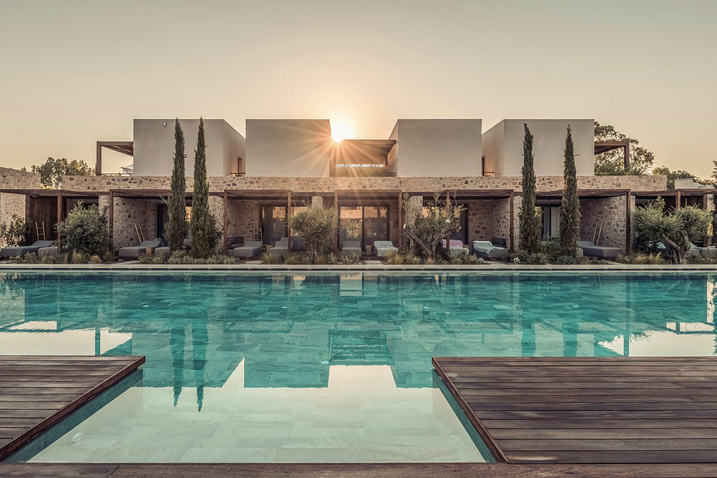 Bild av en modern villa med en pool framför, omgiven av växtlighet och träd, med solens strålar synliga bakom byggnaden. (AI-genererad text)