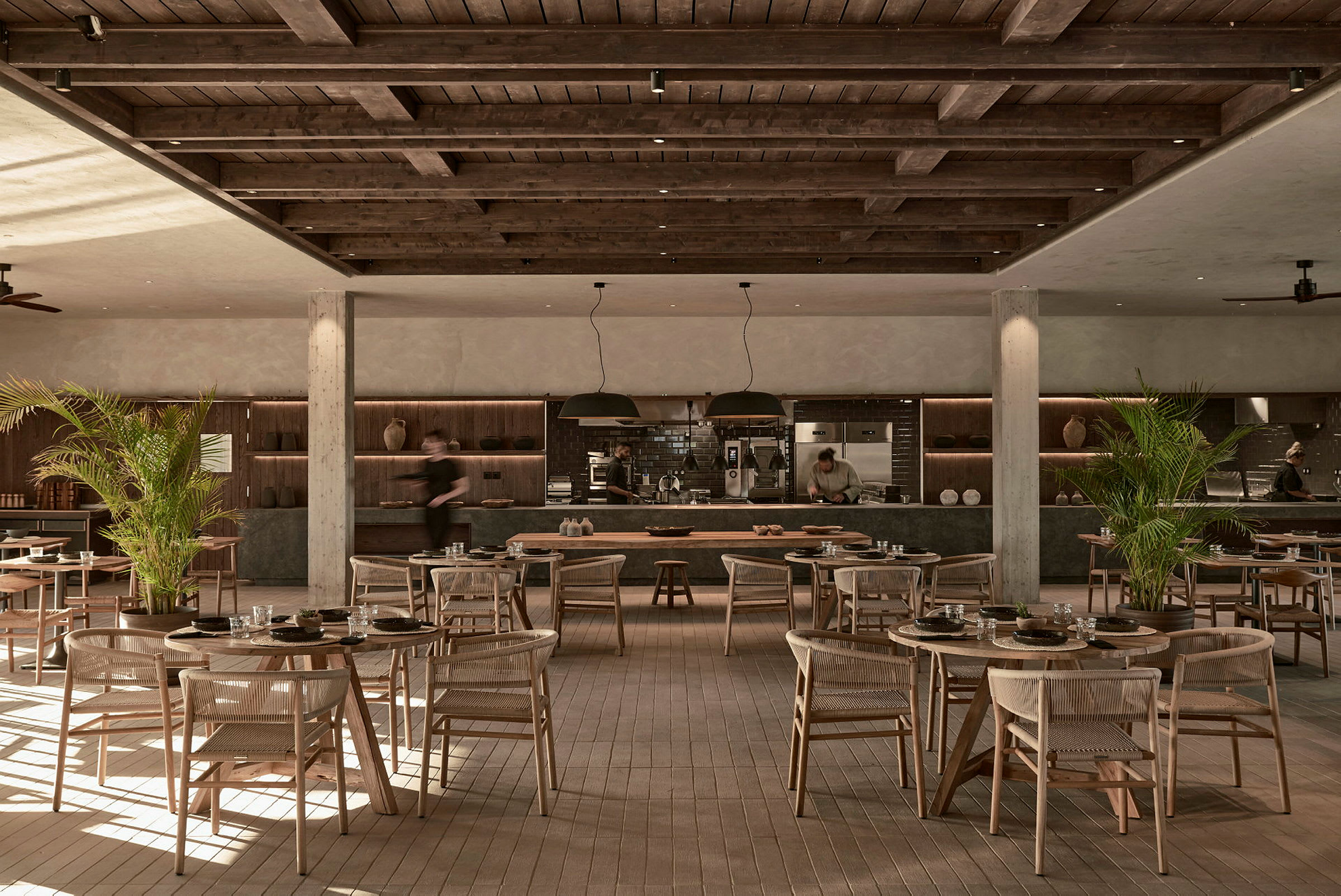 Restaurang med träinredning och bord utspridda över golvet, med öppen köksyta i bakgrunden. (AI-genererad text)