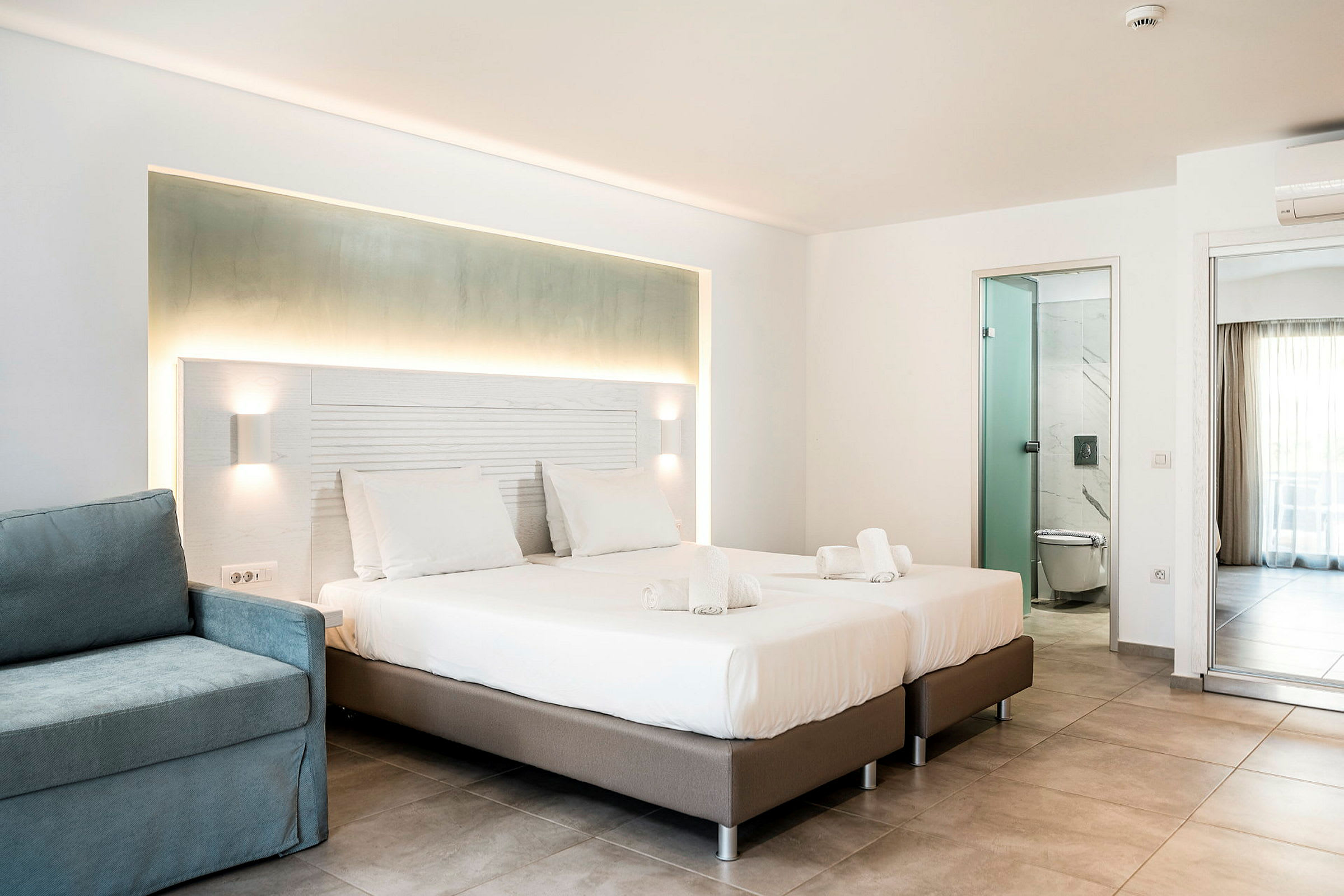 Ett ljust hotellrum med två enkelsängar, en blå fåtölj och ett badrum med glasdörr. (AI-genererad text)