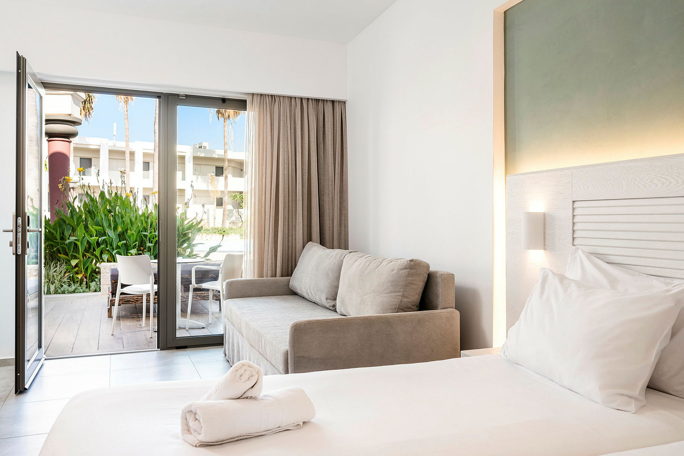 Ett modernt hotellrum med en dubbelsäng, en grå soffa och en öppen glasdörr som leder till en terrass med stolar och grönska. (AI-genererad text)