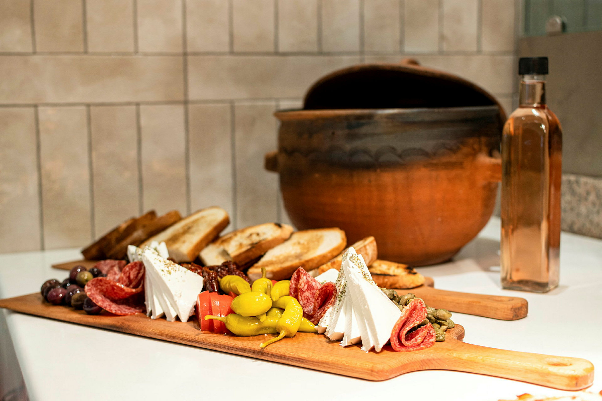 En skärbräda med olika antipasti som ost, salami, oliver och bröd, samt en lergryta och en glasflaska i bakgrunden. (AI-genererad text)