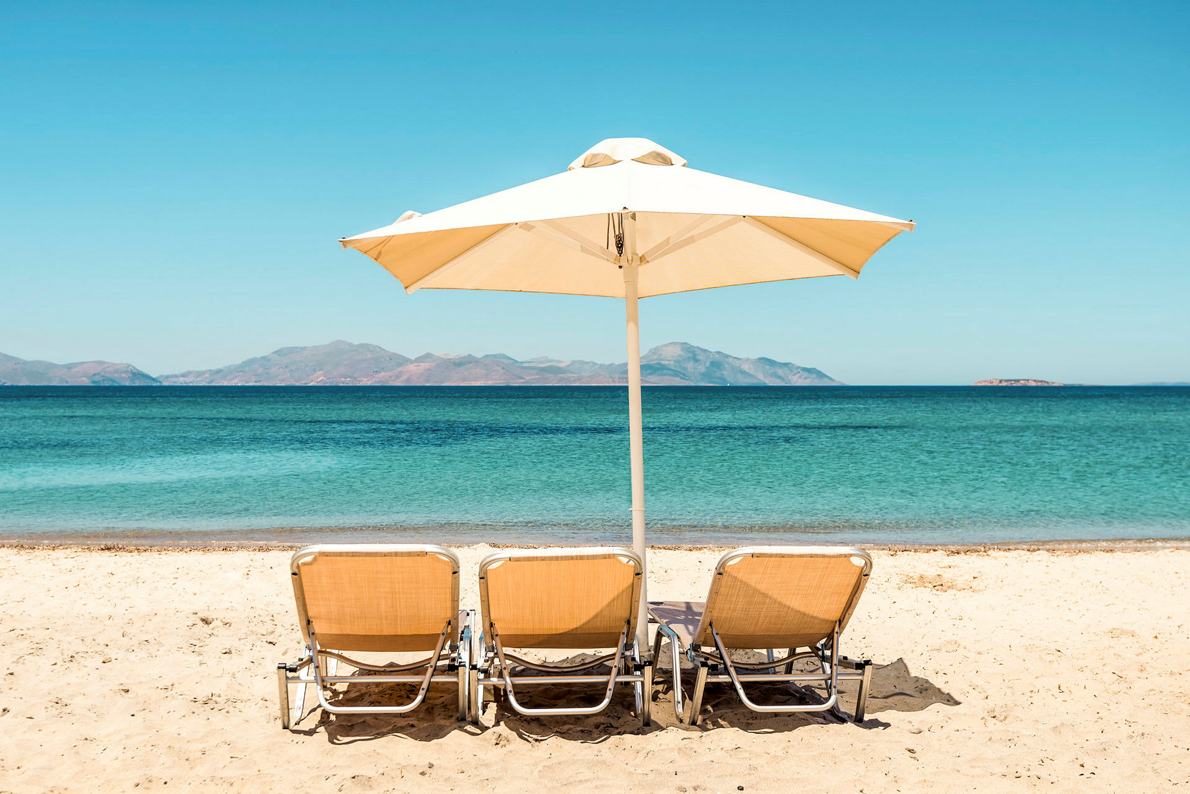 Tre solstolar och ett parasoll står på en sandstrand med utsikt över havet och berg i bakgrunden. (AI-genererad text)