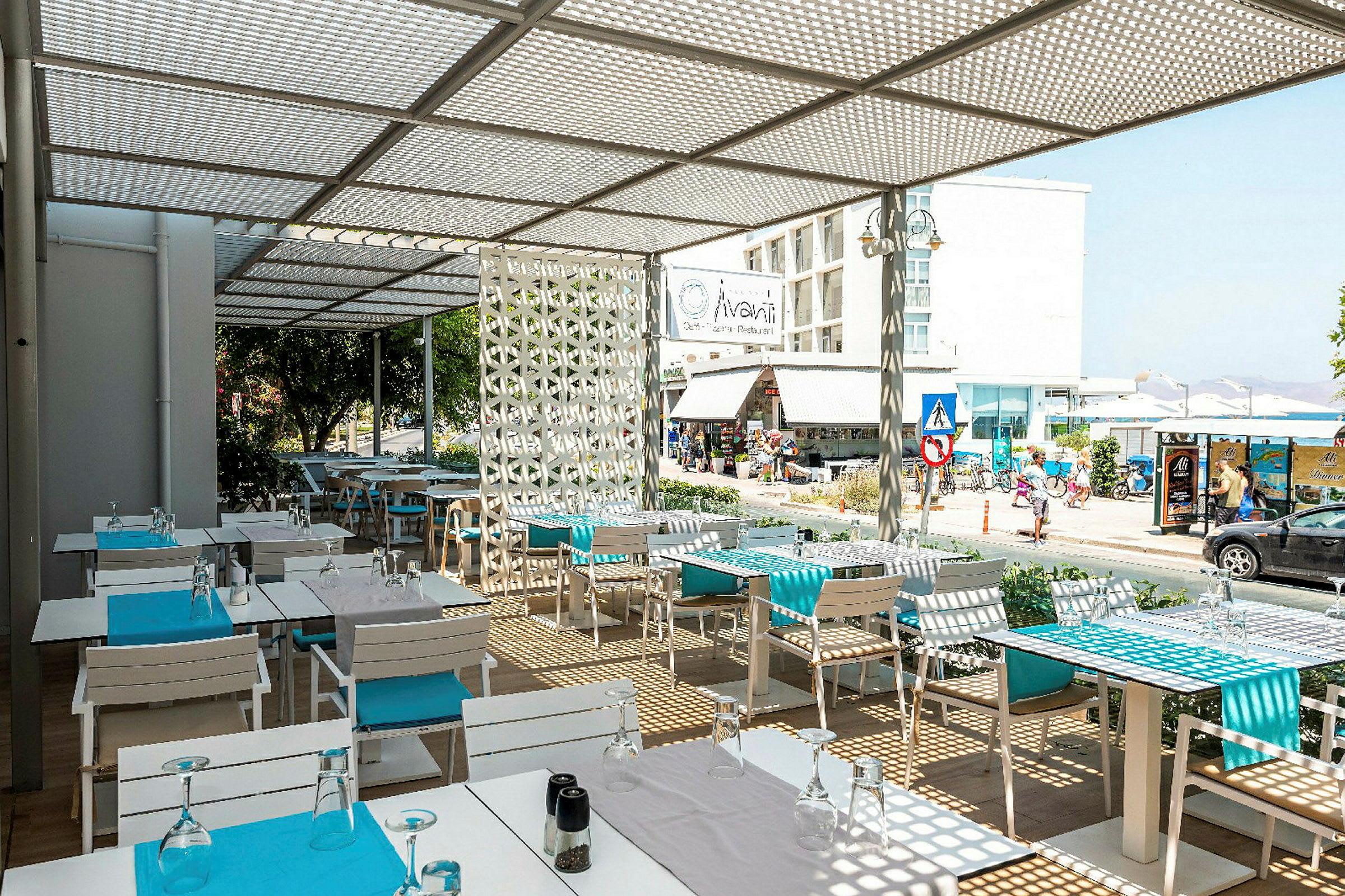 En utomhusrestaurang med vita bord och blå detaljer, med utsikt mot en gata och byggnader. (AI-genererad text)