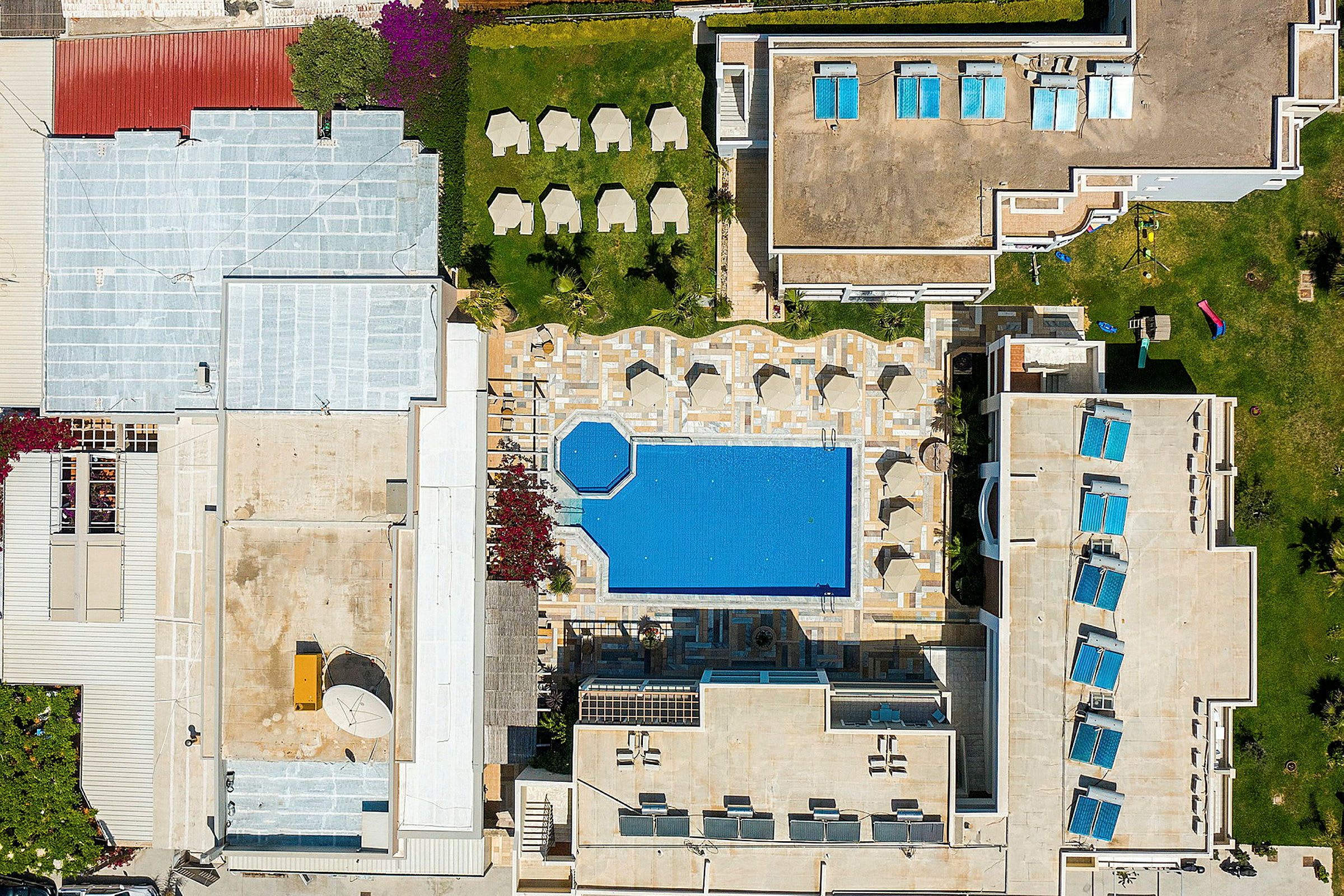 Flygfoto av en hotellpool omgiven av solstolar och byggnader. (AI-genererad text)