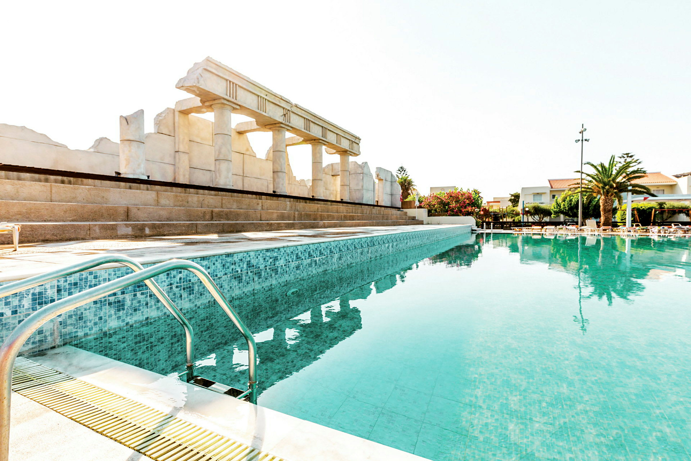 En ljusblå pool med antika ruiner och byggnader i bakgrunden. (AI-genererad text)