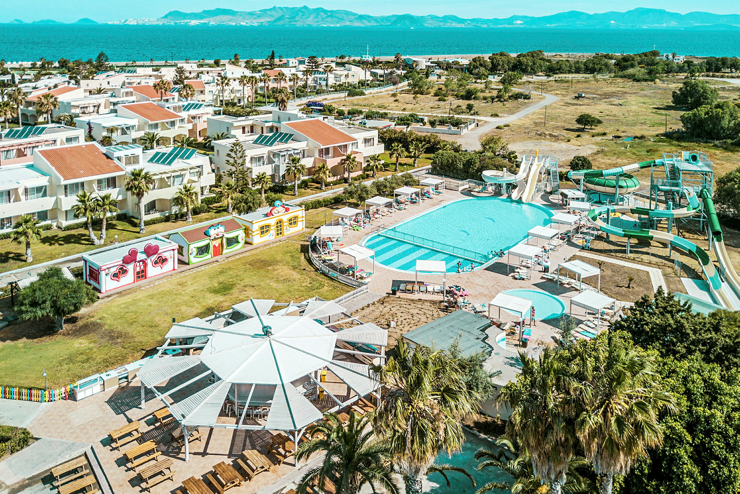 Flygfoto av ett hotellområde med pooler och vattenrutschbanor, omgiven av byggnader och grönska nära havet. (AI-genererad text)