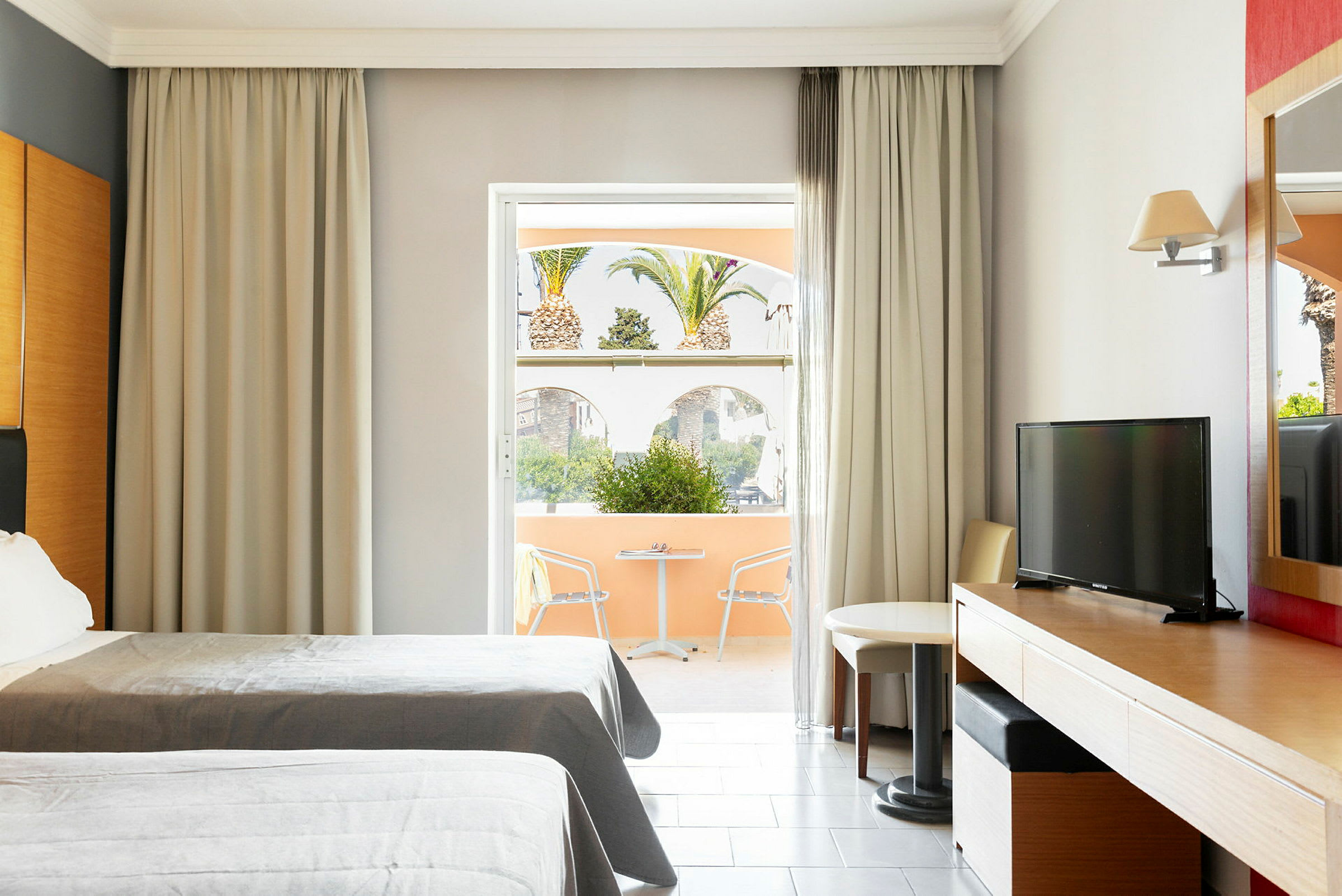 Hotellrum med två sängar, skrivbord, TV och en sittgrupp vid ett fönster ut mot en terrass med utsikt över palmer. (AI-genererad text)