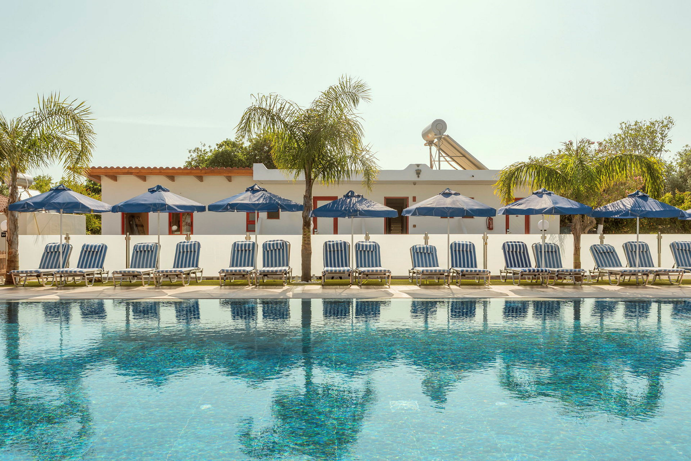 Utsikt över en pool med solstolar och parasoll på rad framför en vit byggnad. (AI-genererad text)