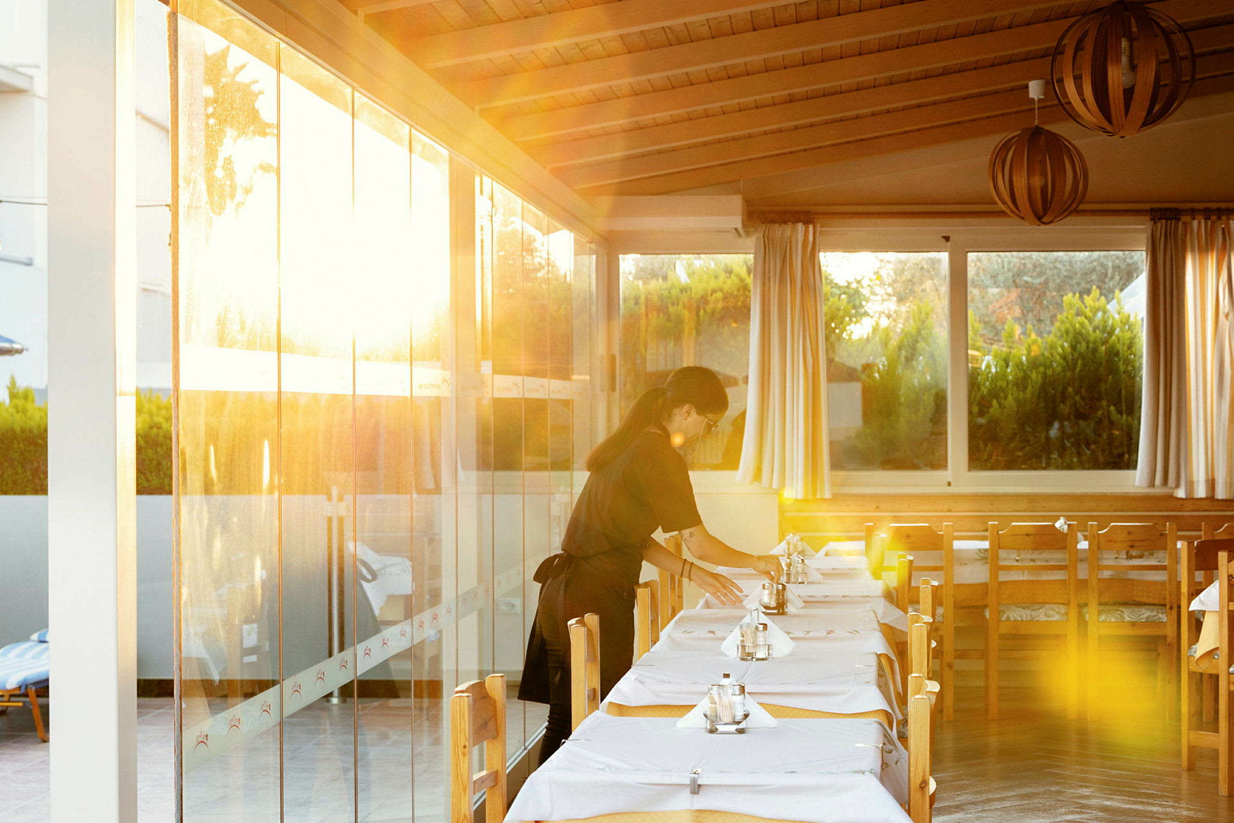 En restaurang med uppdukade bord och solsken som lyser genom glasväggarna, krukväxter syns utanför fönstret. (AI-genererad text)