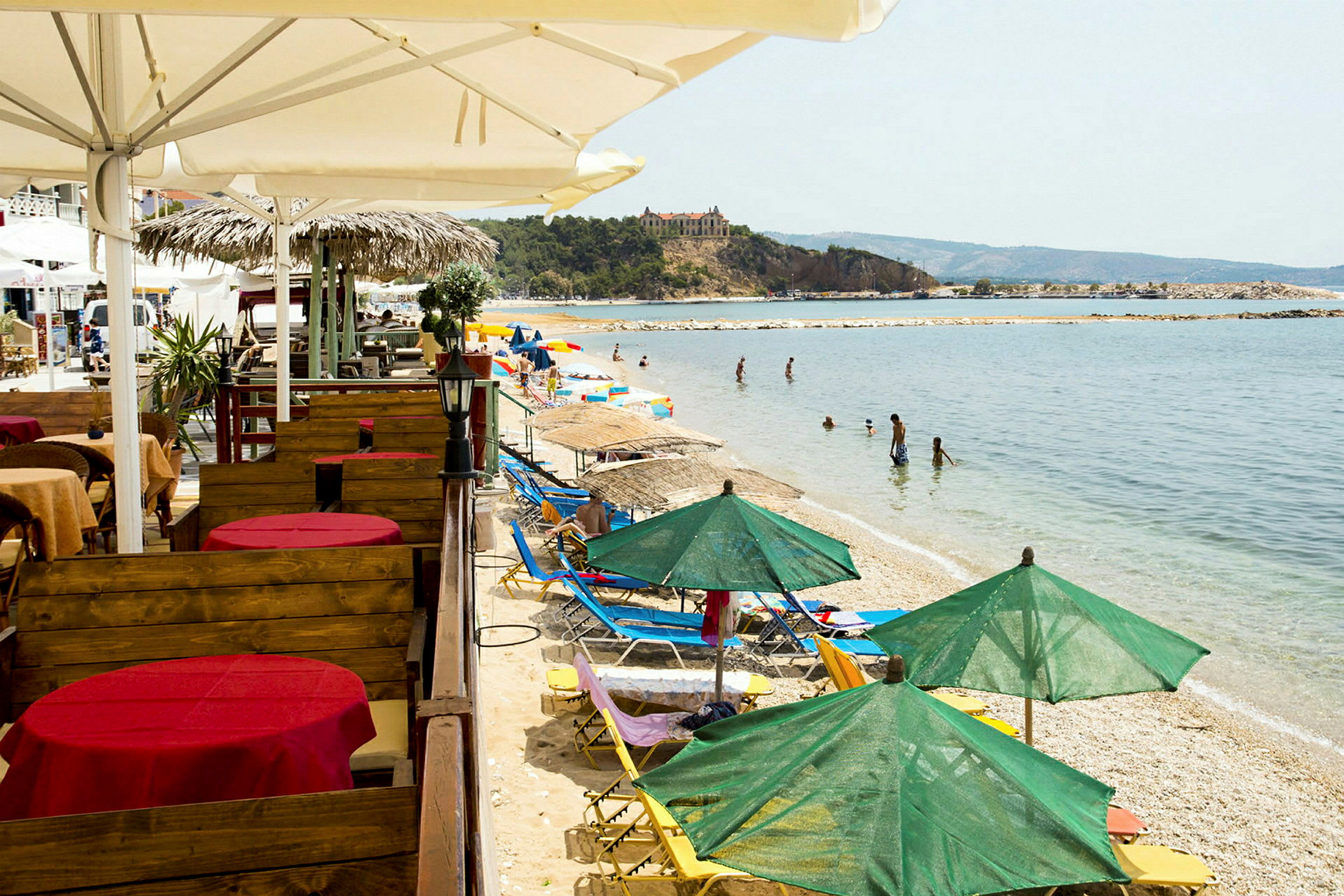 Strand med solstolar och gröna parasoller samt bord med röda dukar vid en restaurang. (AI-genererad text)