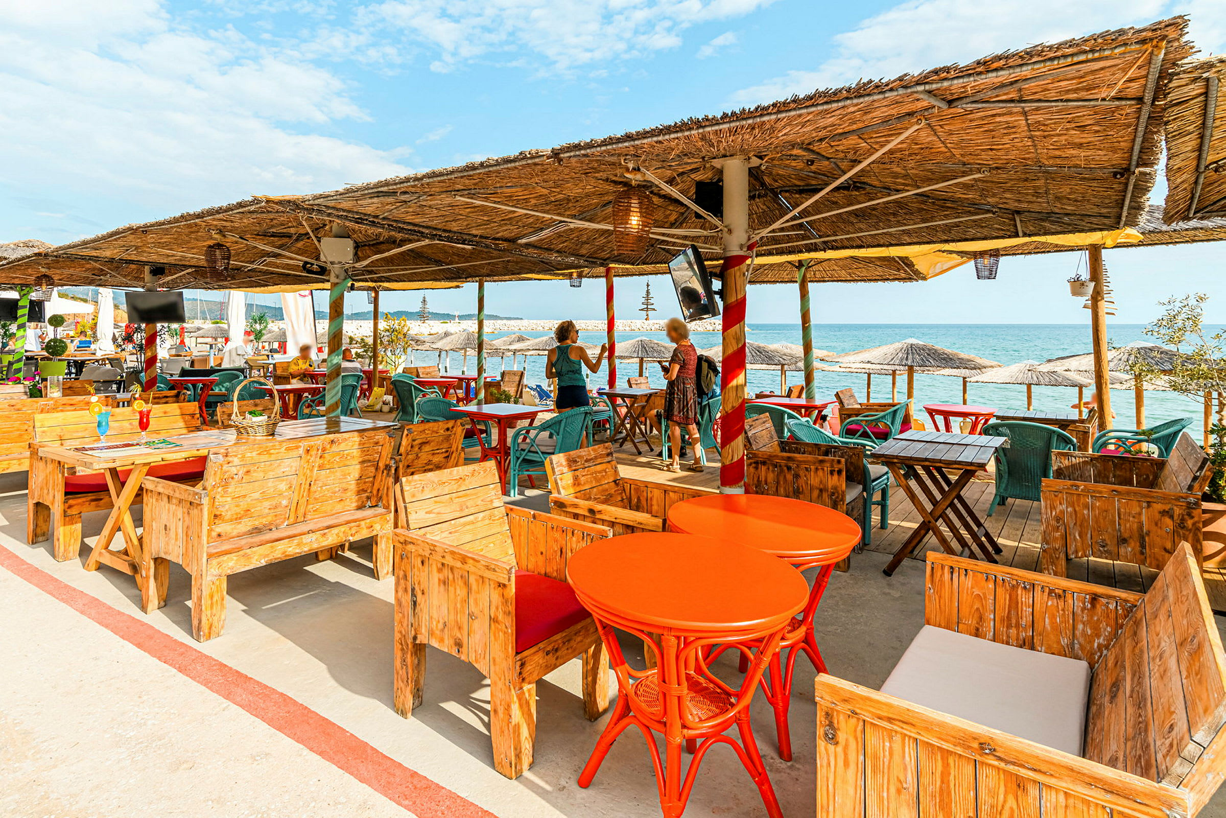 En utomhusrestaurang vid havet med trästolar, orange bord, och halmtak. I bakgrunden syns parasoller och havet. (AI-genererad text)