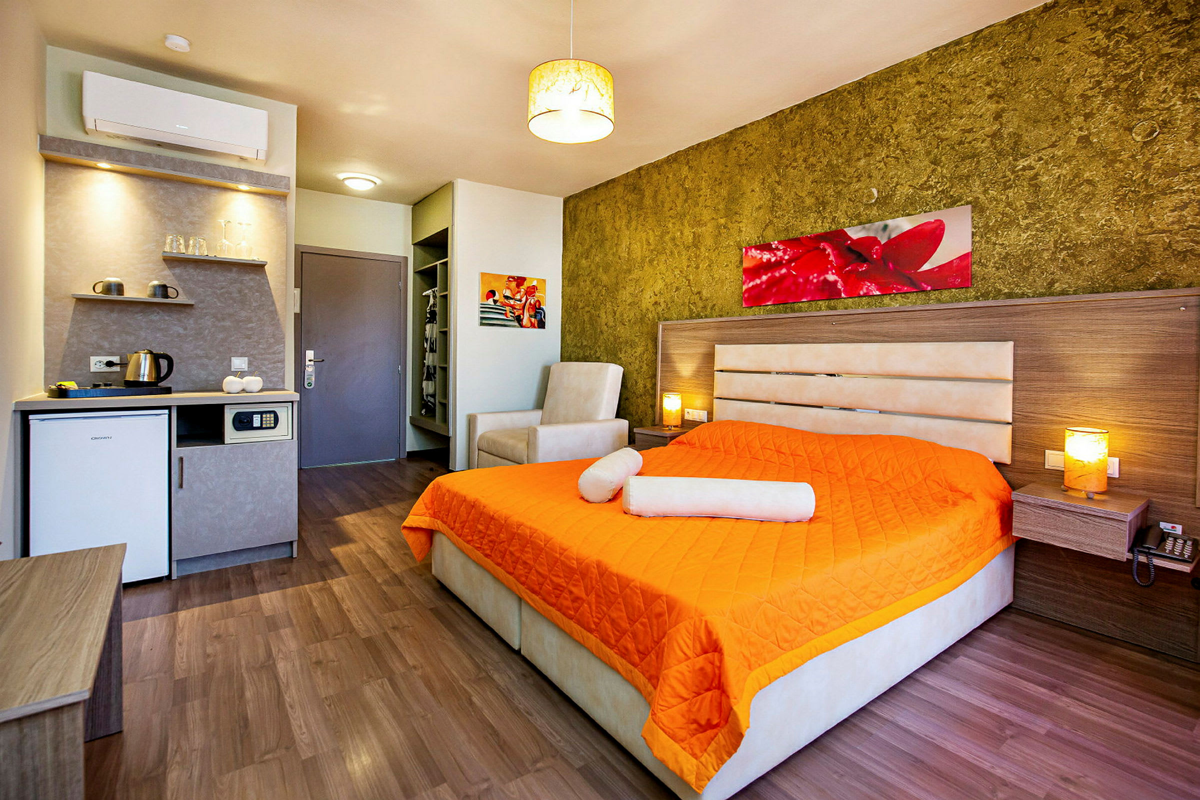 Ett hotellrum med en säng täckt med orange överkast, en fåtölj och ett litet köksutrymme. (AI-genererad text)