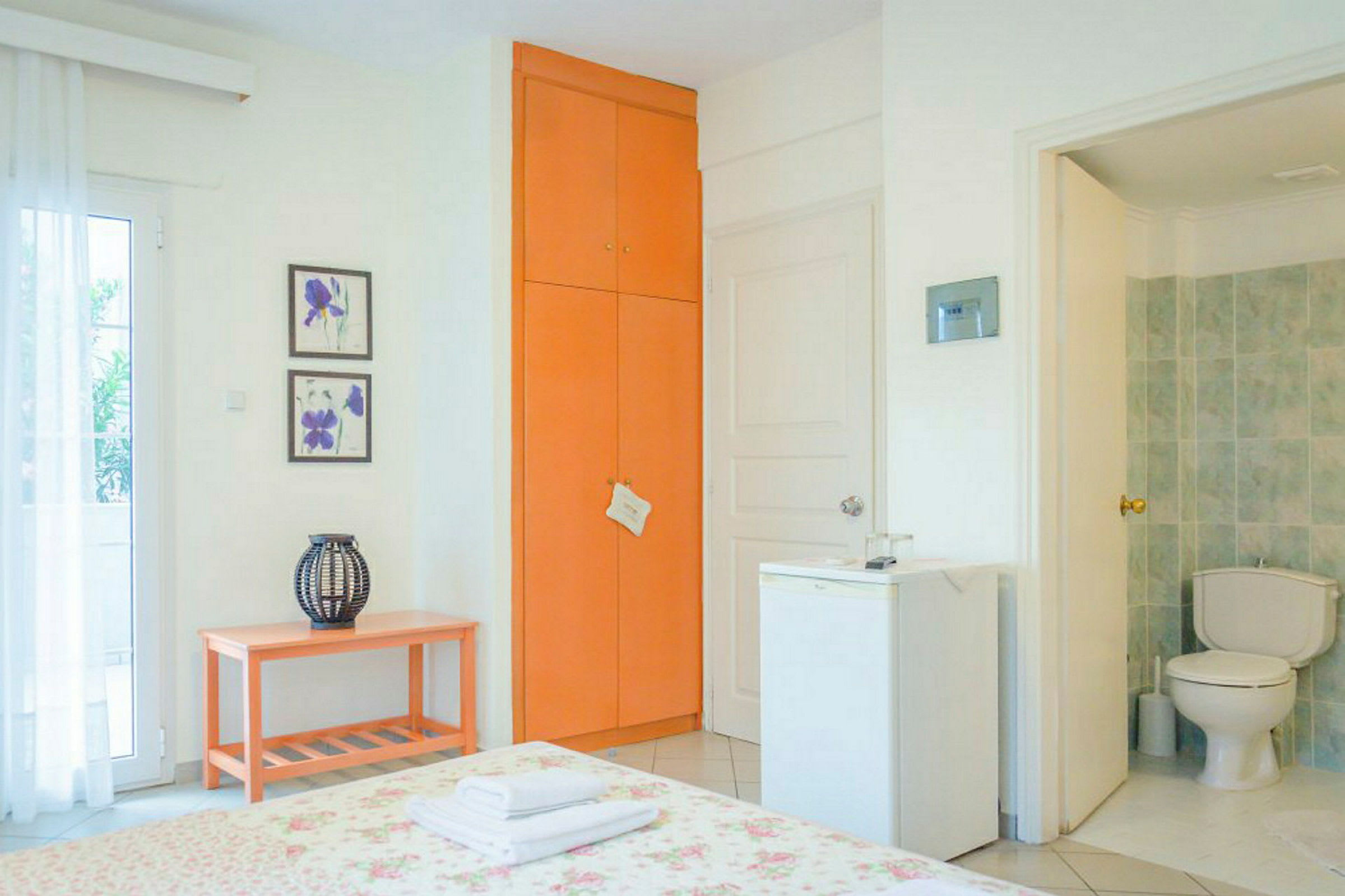 Ett hotellrum med orange skåp, en säng med blommigt överkast och en minikyl bredvid en dörr till badrummet. (AI-genererad text)