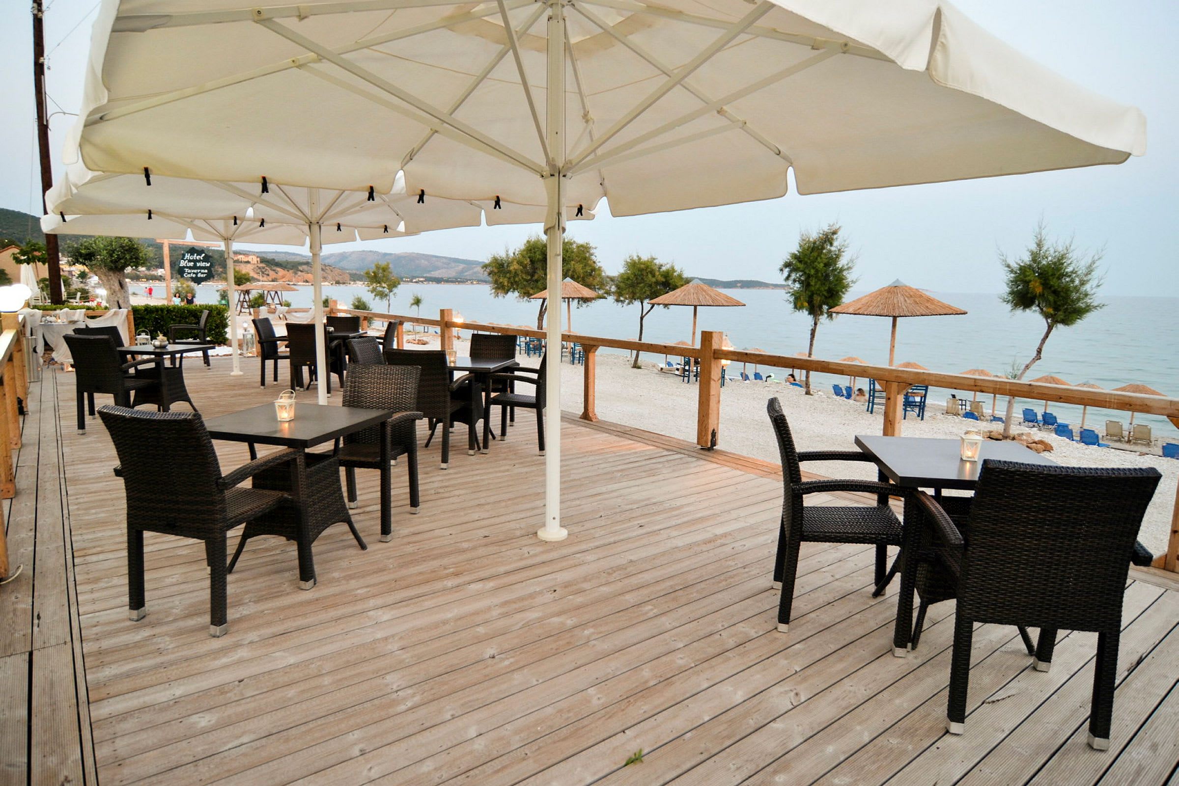 En träveranda med möblerade bord och stolar samt parasoller, med utsikt mot stranden och havet. (AI-genererad text)
