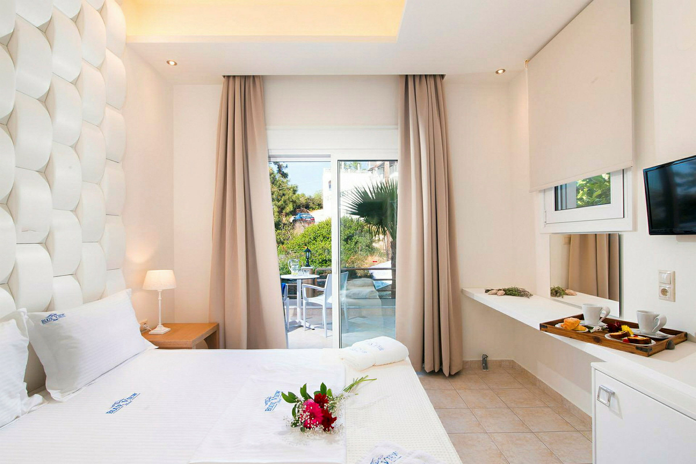 Hotellrum med vit säng, dekorationer och utsikt mot en grönskande terrass genom stora fönster. (AI-genererad text)