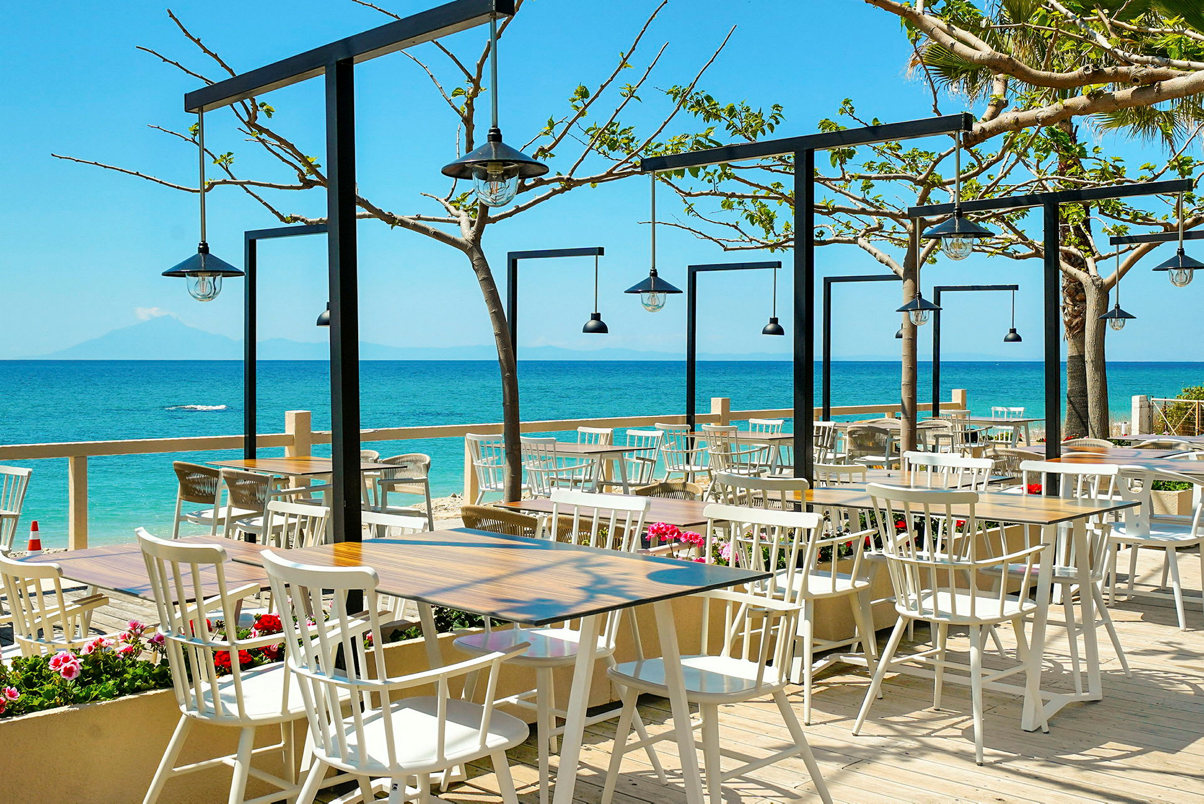 Utomhusrestaurang med stolar och bord vid havet. Blå himmel och blommor runt om. (AI-genererad text)