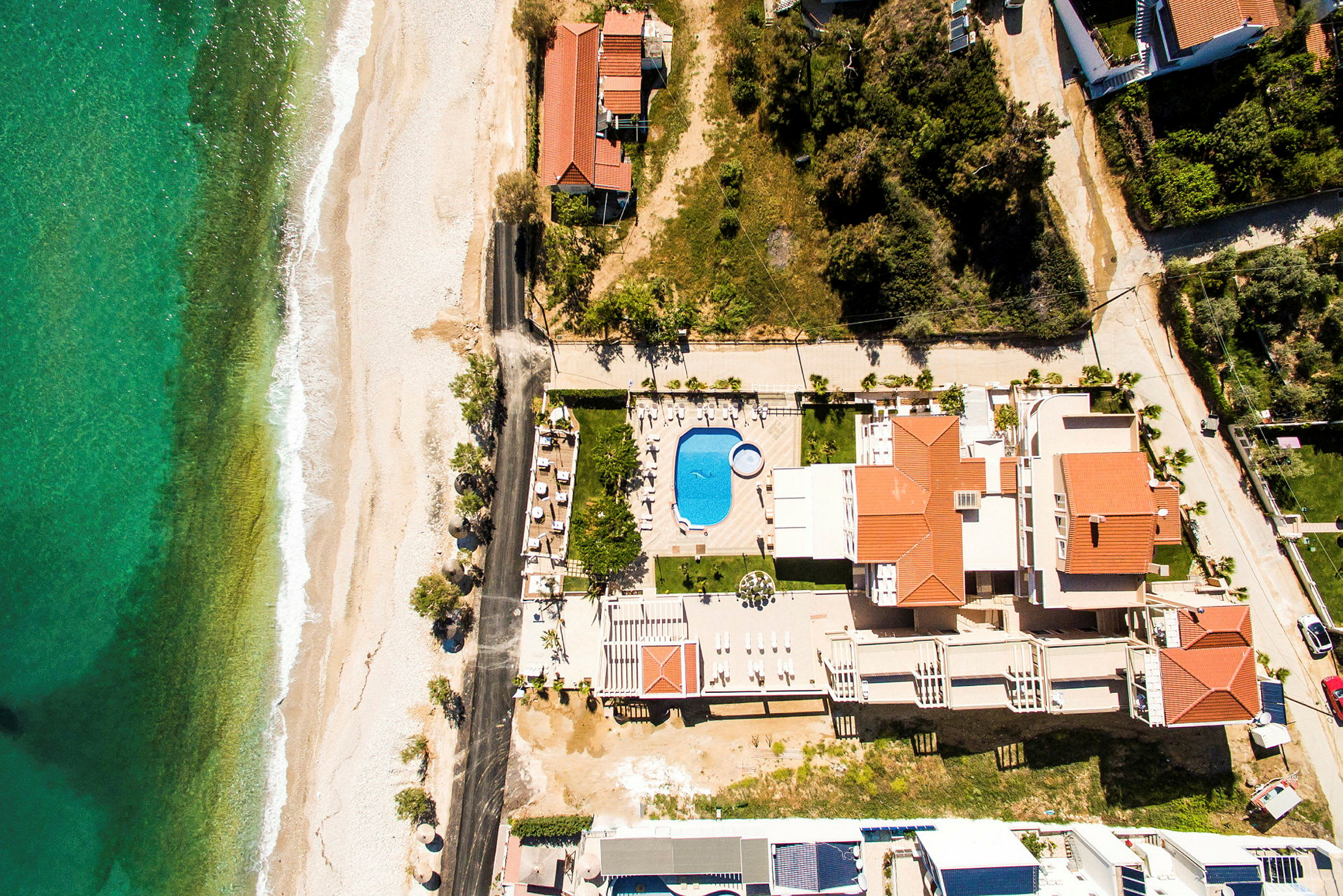 Flygfoto av en anläggning med flera byggnader med röda tak, en pool och närliggande strand. (AI-genererad text)