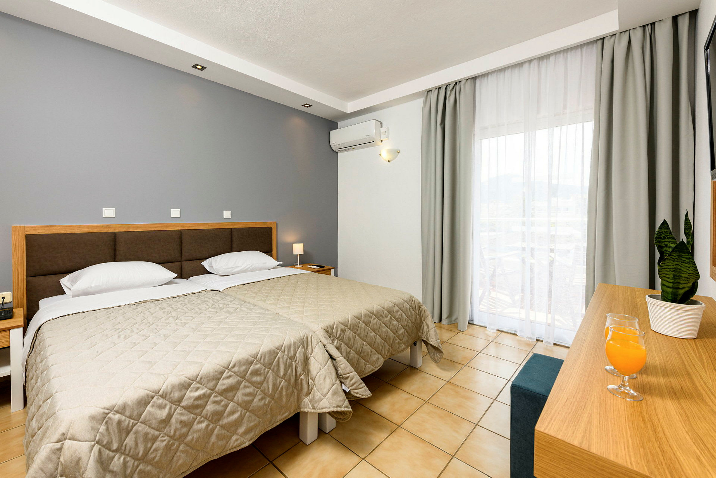 Hotellrum med två enkelsängar, ett skrivbord med en växt och dryck, och stora fönster med ljusa gardiner. (AI-genererad text)