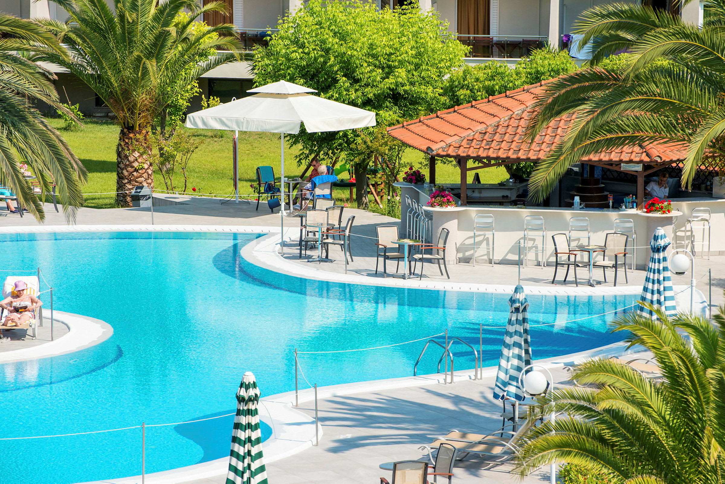 En utomhuspool med omgivande solstolar, parasoller och en poolbar under ett tak med röda takpannor. (AI-genererad text)