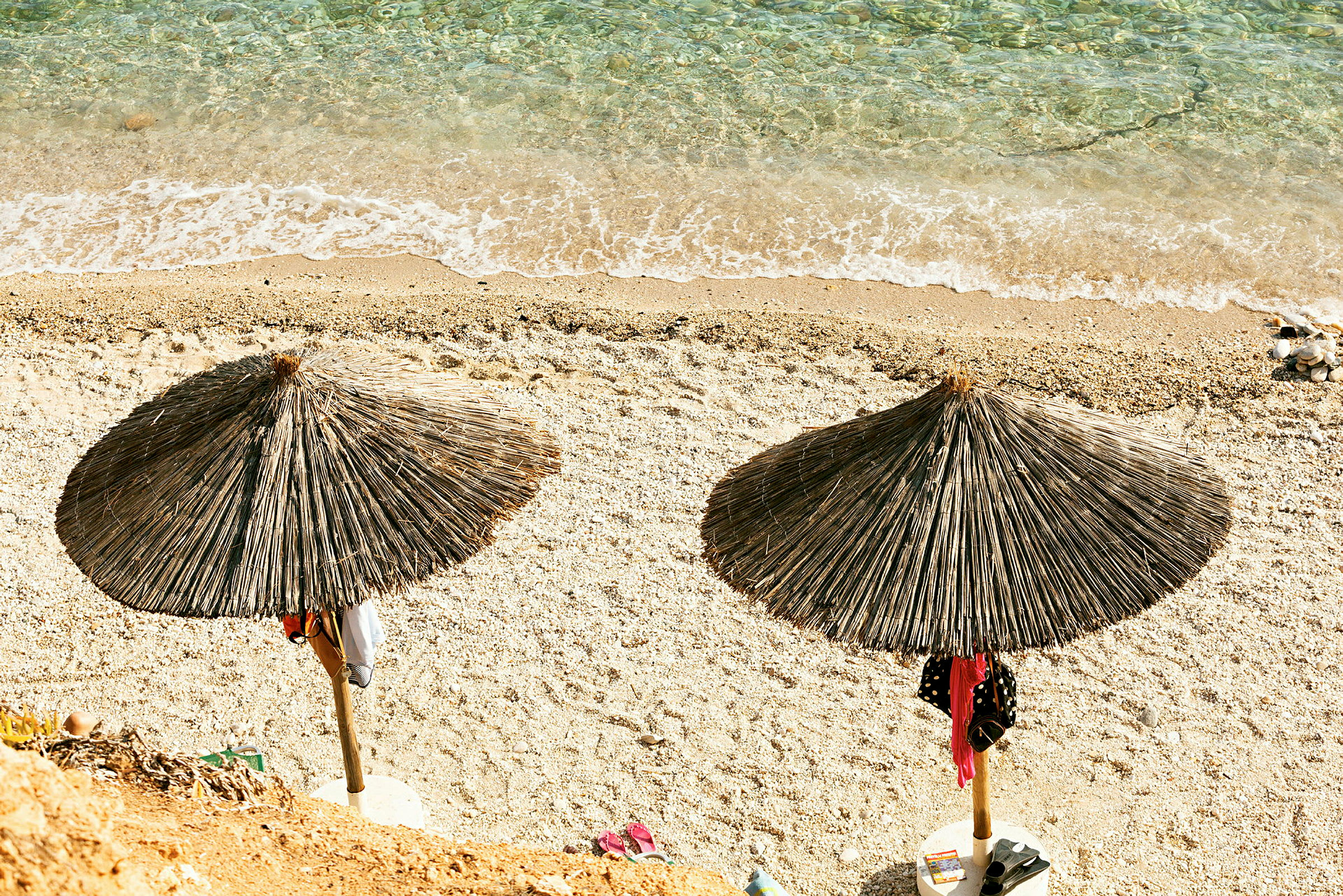 Två halmtakparaplyer står på en stenig strand nära vattnet. (AI-genererad text)