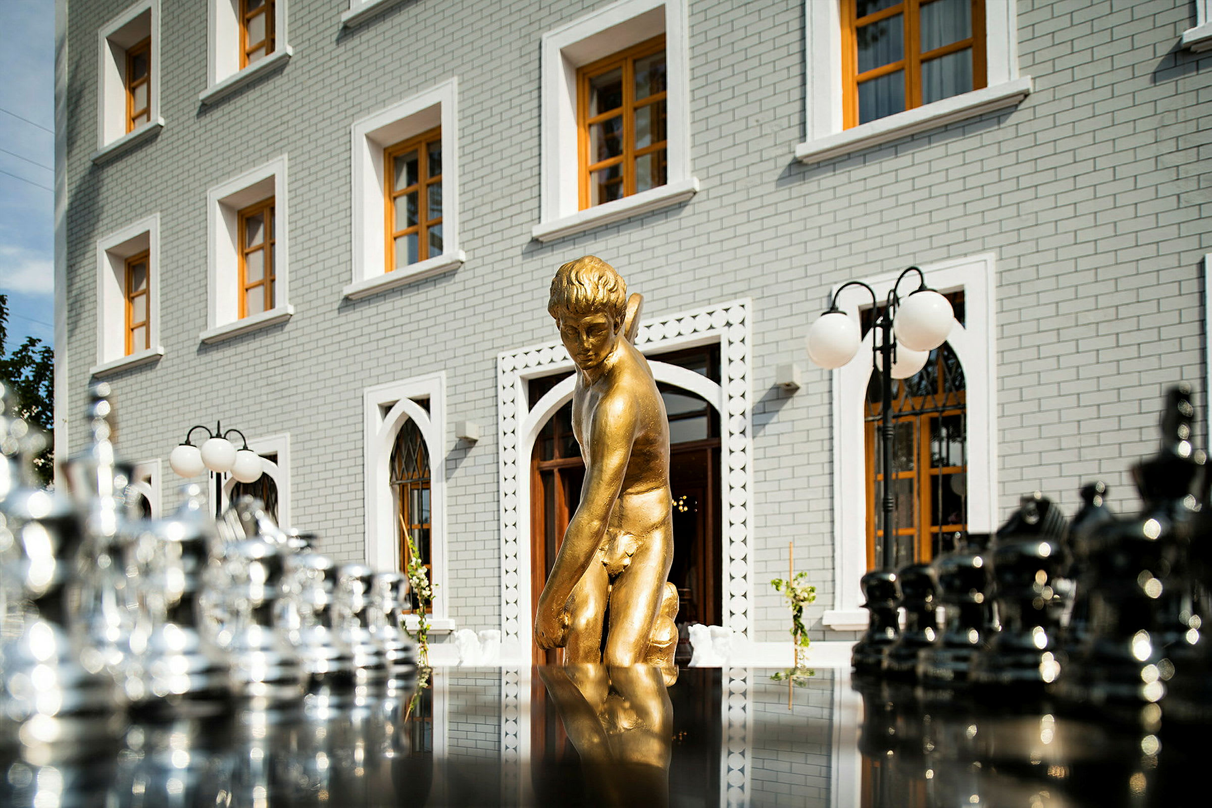 En guldfärgad staty framför en byggnad med vita tegelväggar och stora fönster. Chesspjäser ses i förgrunden. (AI-genererad text)