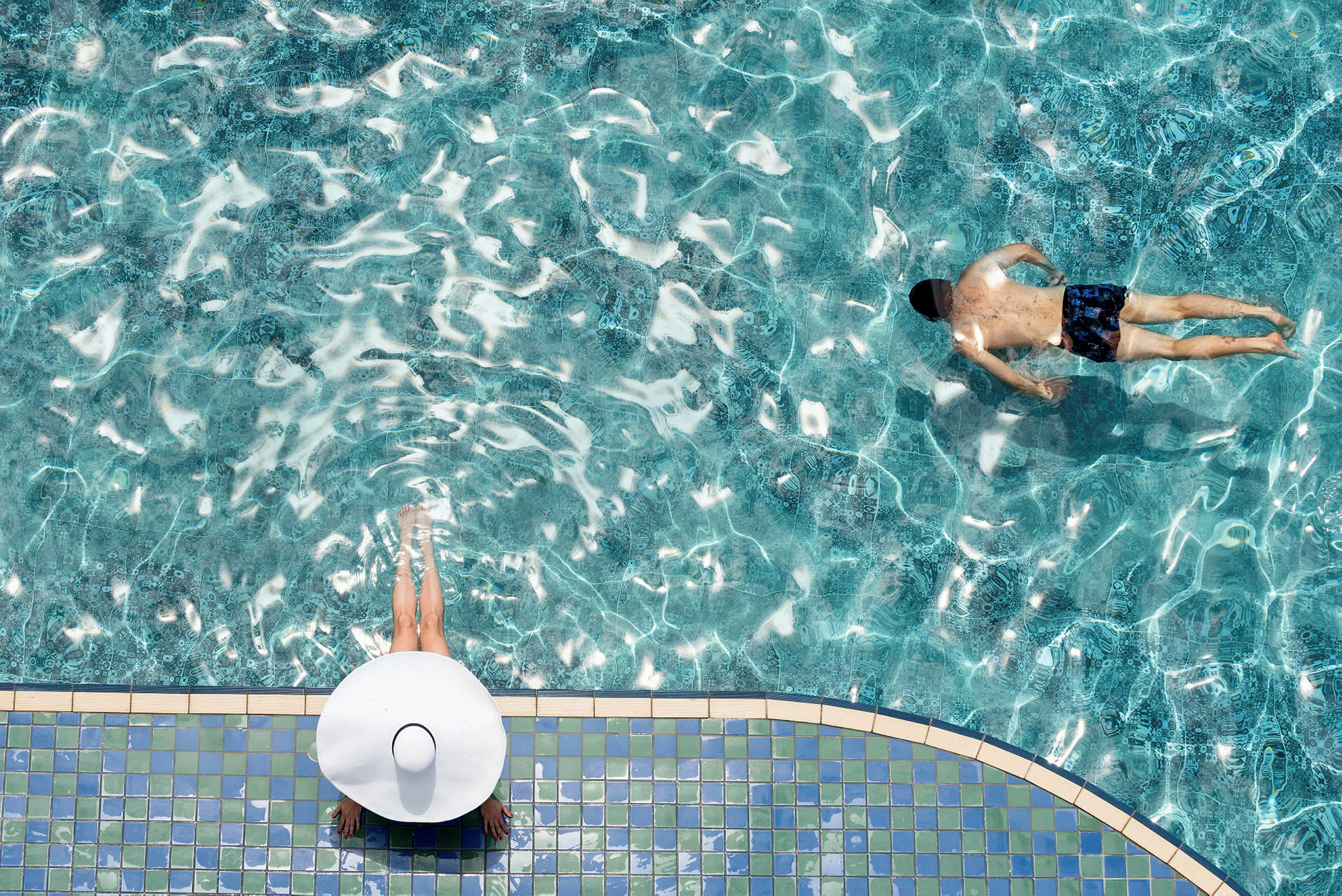 Person med vit hatt sitter vid kanten av en pool, medan en annan person simmar i det klara vattnet. (AI-genererad text)