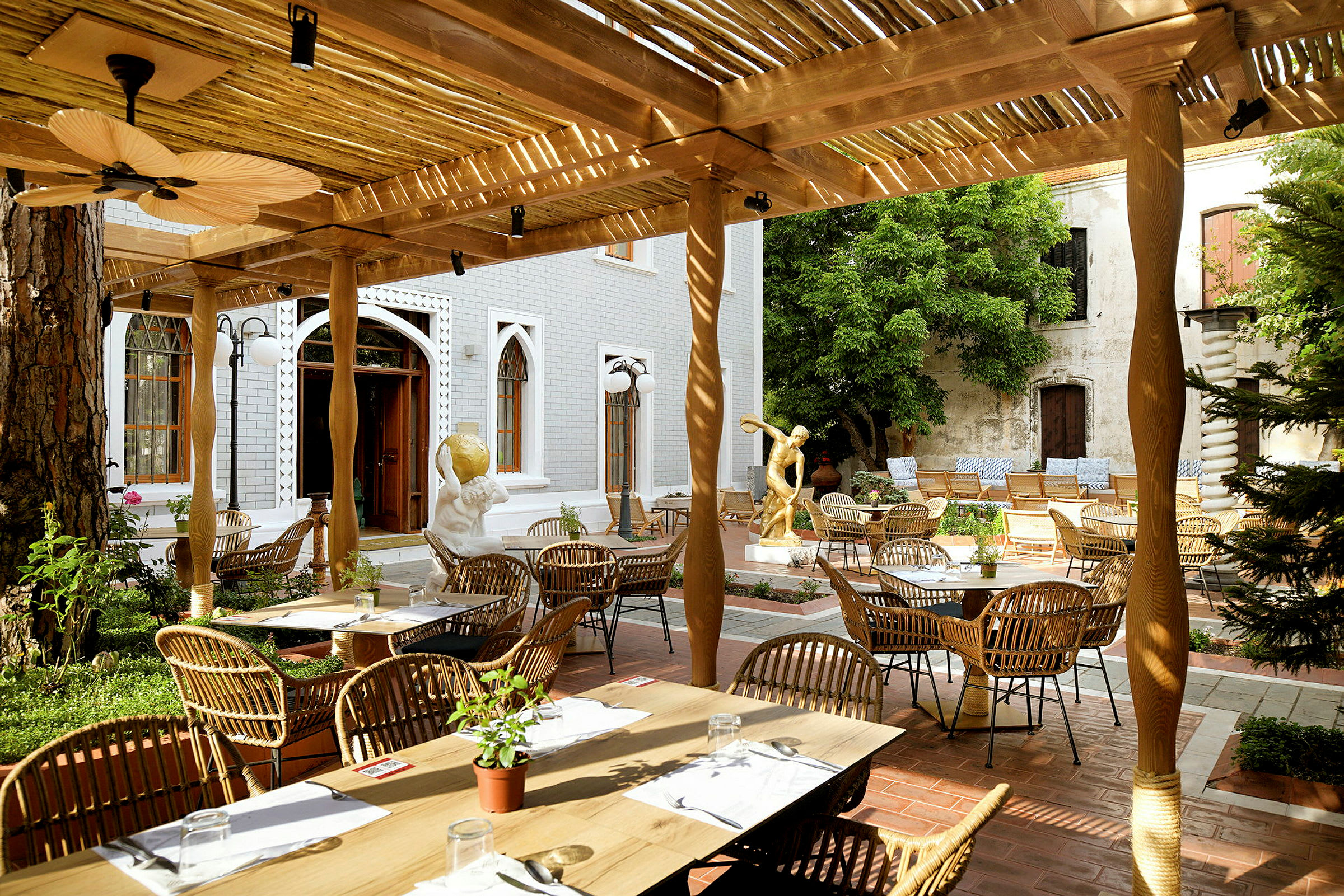 En utomhus restaurang med bord och stolar under en träpergola, omgiven av skulpturer och grönska. (AI-genererad text)